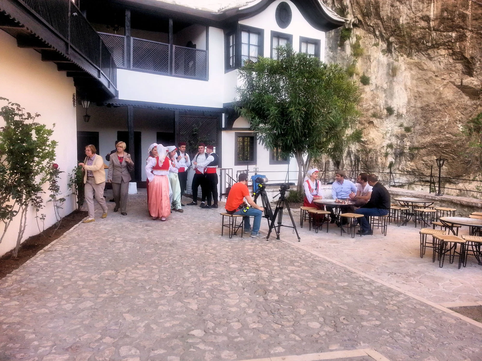 A group of people dressed in traditional costumes, some appear to be preparing or performing a cultural dance or celebration outside a white building with black accents, next to a rocky cliff, with tables and chairs nearby.