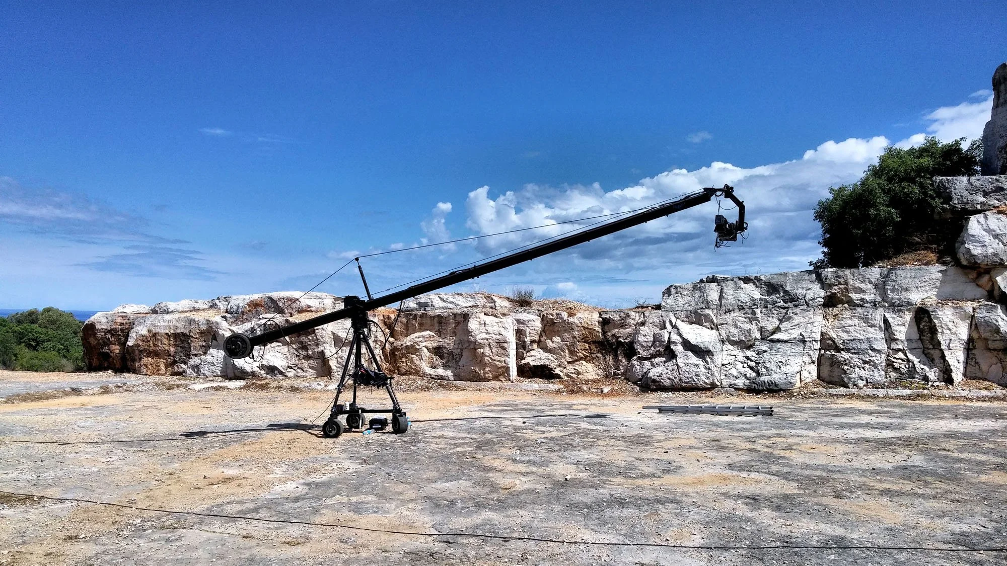 A camera crane set up on rocky terrain outdoors with a blue sky and scattered clouds in the background.
