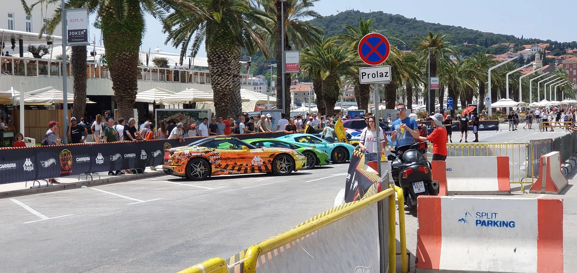 Multiple colorful sports cars lined up on a street during a car event, with people walking and gathered around, palm trees, buildings, and mountains in the background.