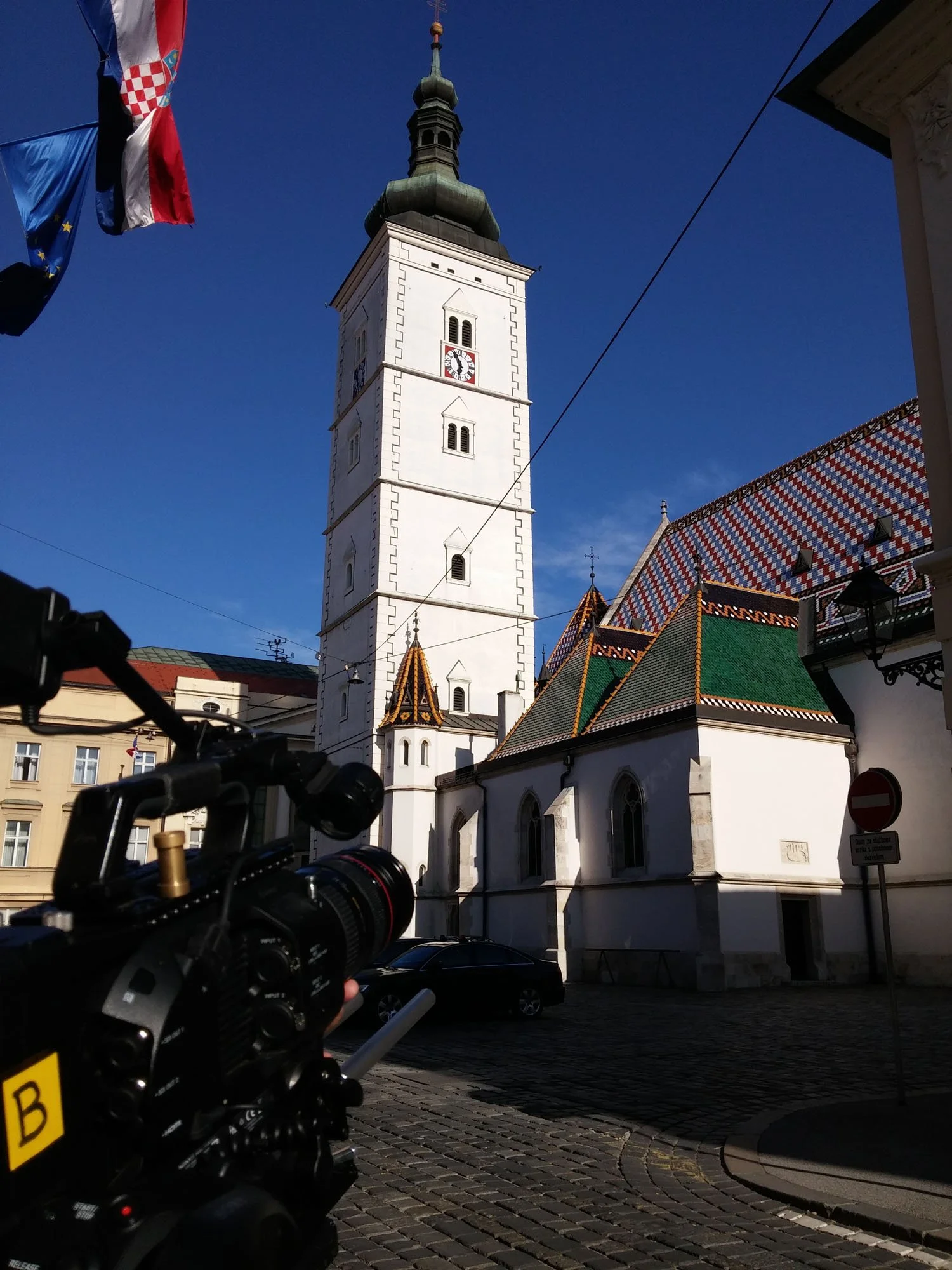 Photo of a tall white clock tower with a dark green roof and smaller turrets, set against a clear blue sky. Flags are visible on the left side, and the foreground shows a camera on a tripod and parked cars on cobblestone streets.