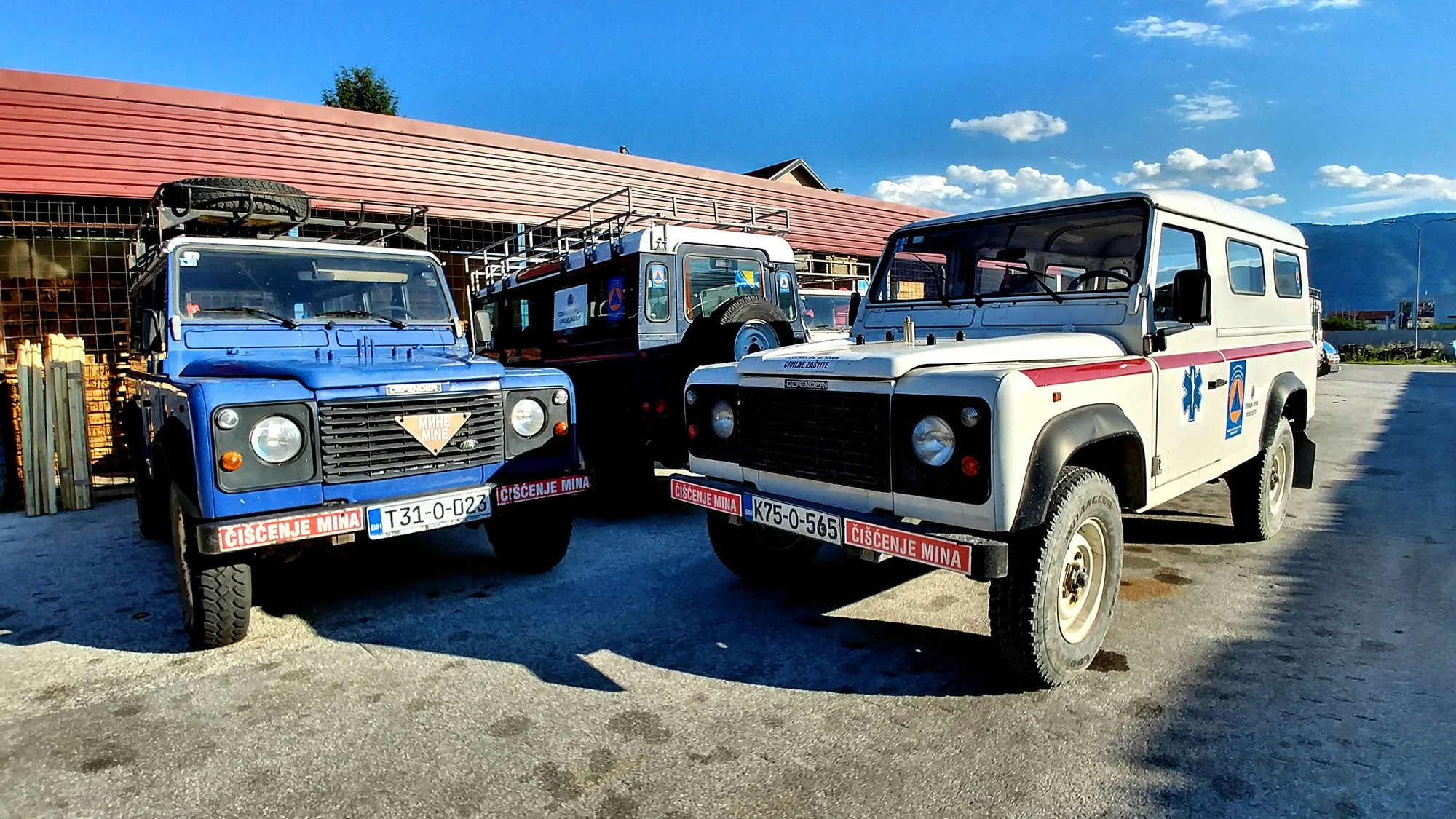Three emergency vehicles parked outside a building, with blue skies overhead. One blue and one white vehicle are in the foreground, both with emergency medical symbols and red warnings on their bumpers. A third, smaller black vehicle is behind them.