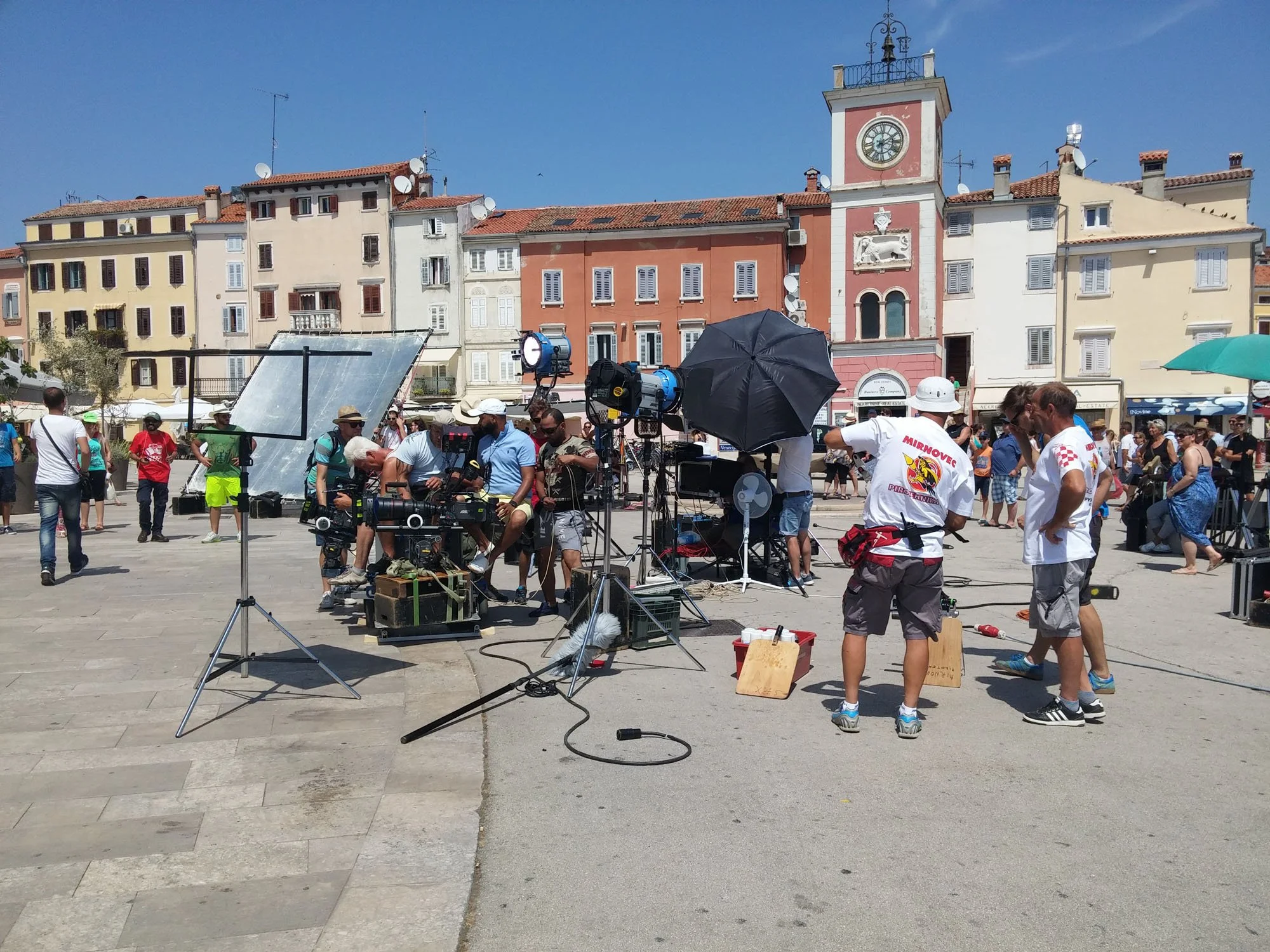 Film set crew working outside in a square with a historic clock tower and colorful buildings in the background. Equipment including cameras, umbrellas, and lighting are visible.