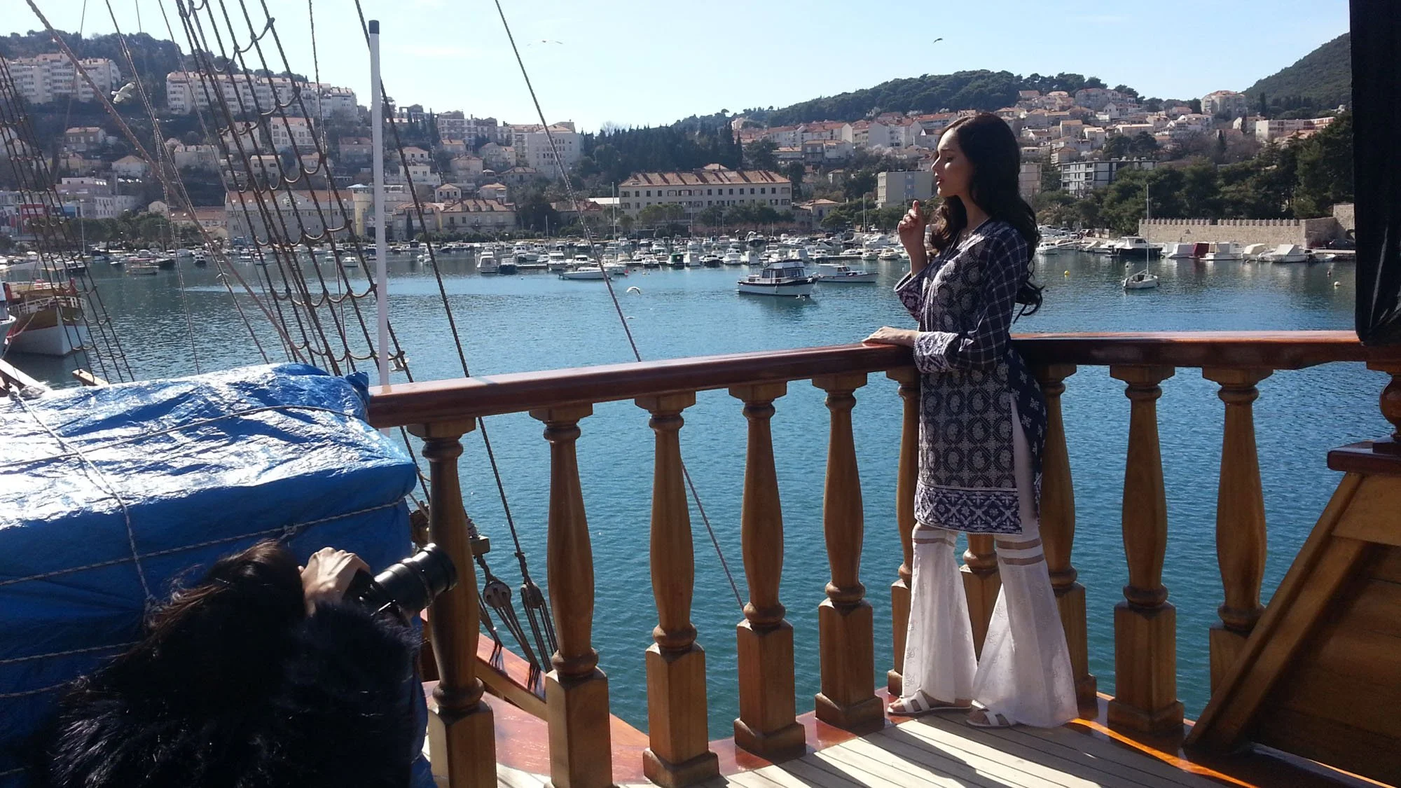 A woman in a patterned dress standing on a ship's deck, looking out at a harbor filled with boats, with a hillside town in the background.