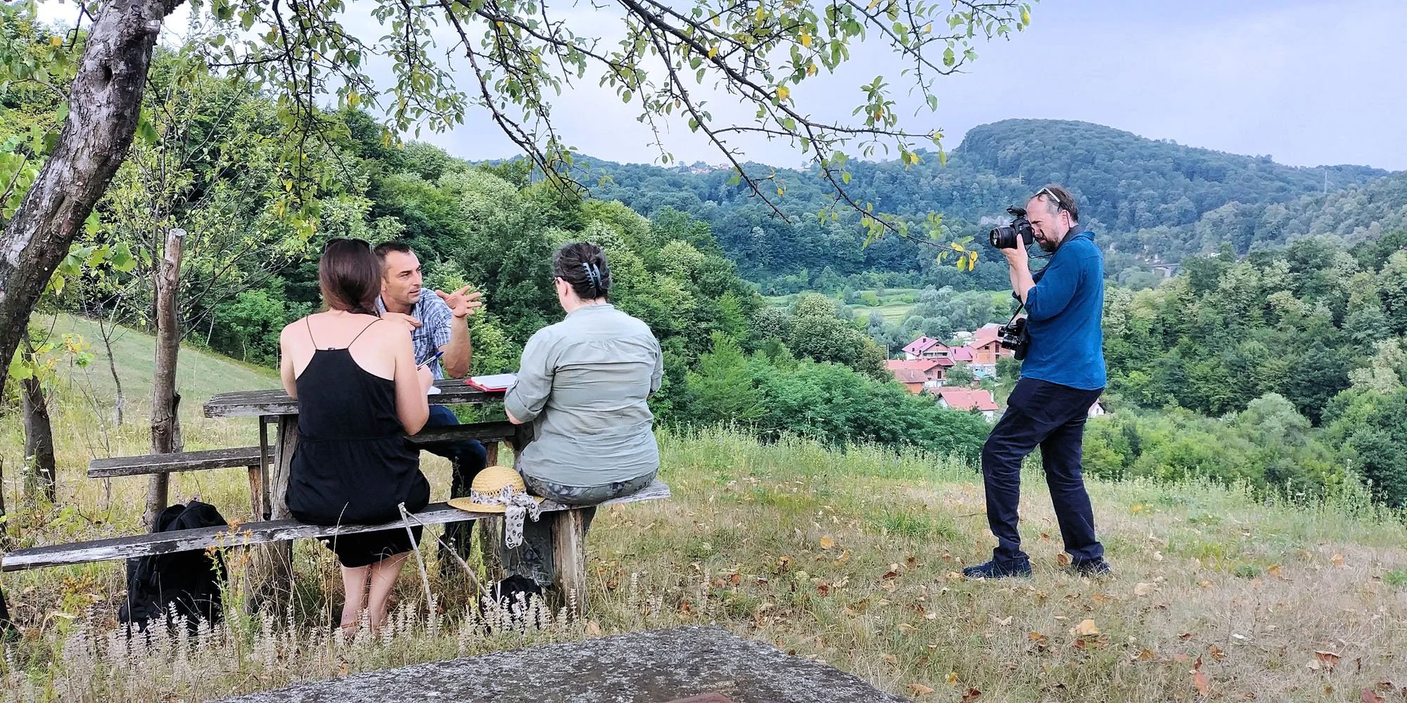 Four people are outdoors in a hilly, wooded area. Three are sitting at a wooden picnic table, engaged in a conversation or interview, while the fourth person, a man, is standing and taking a photograph of them with a camera. The background features g