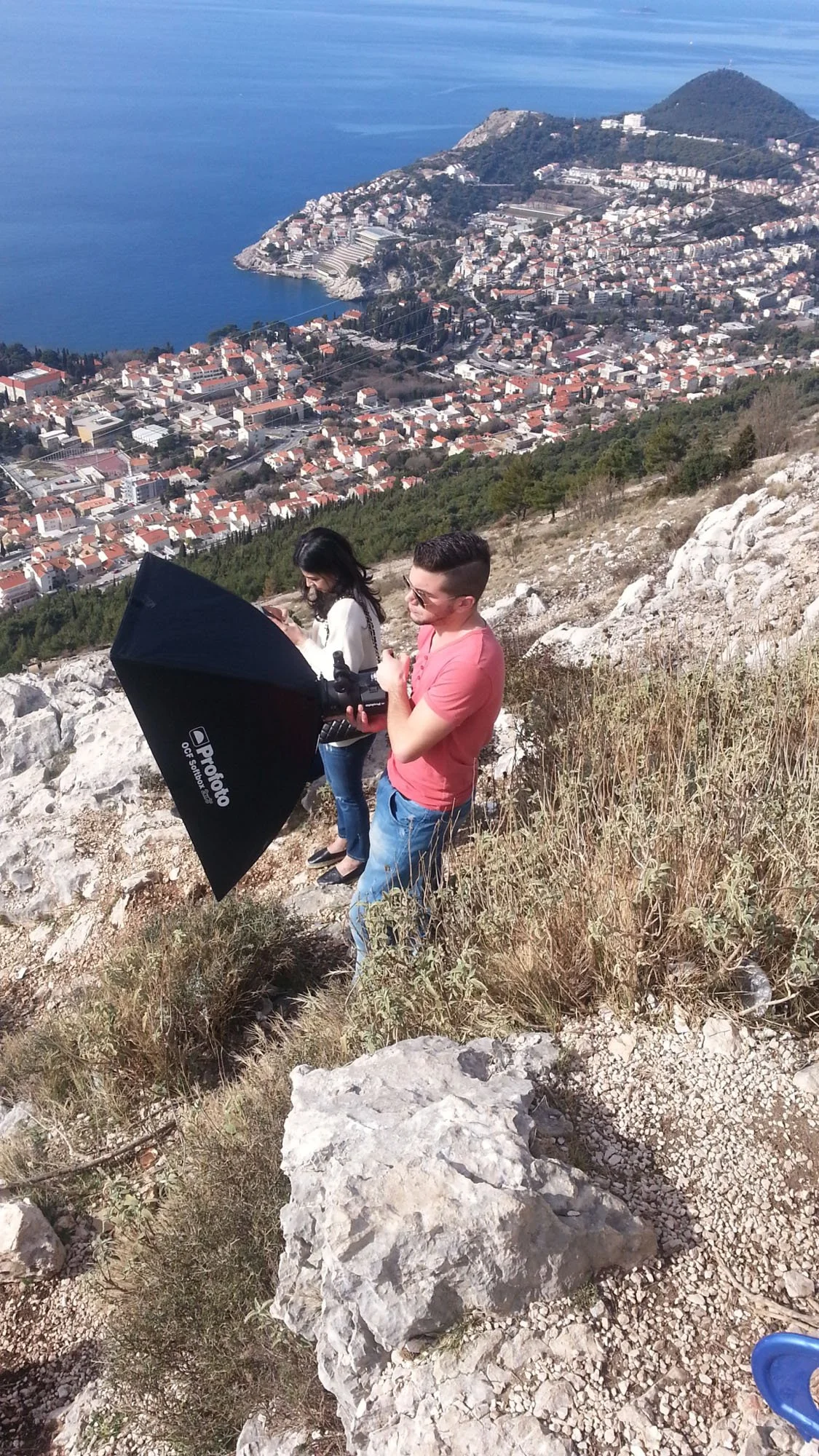 Two people standing on a rocky hillside with a cityscape and water in the background, one holding a large camera reflector.