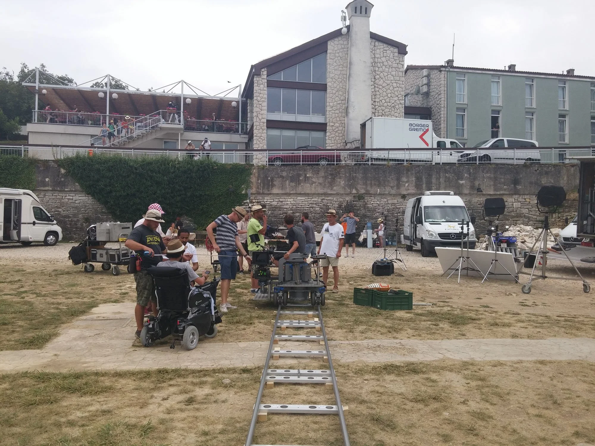 Filming crew setting up equipment on an outdoor set with a building in the background and people working and walking around.