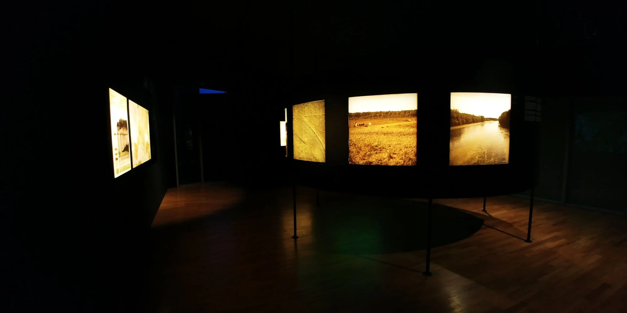 Dimly lit museum display with landscape photographs on illuminated screens, showcasing fields, trees, and a river.