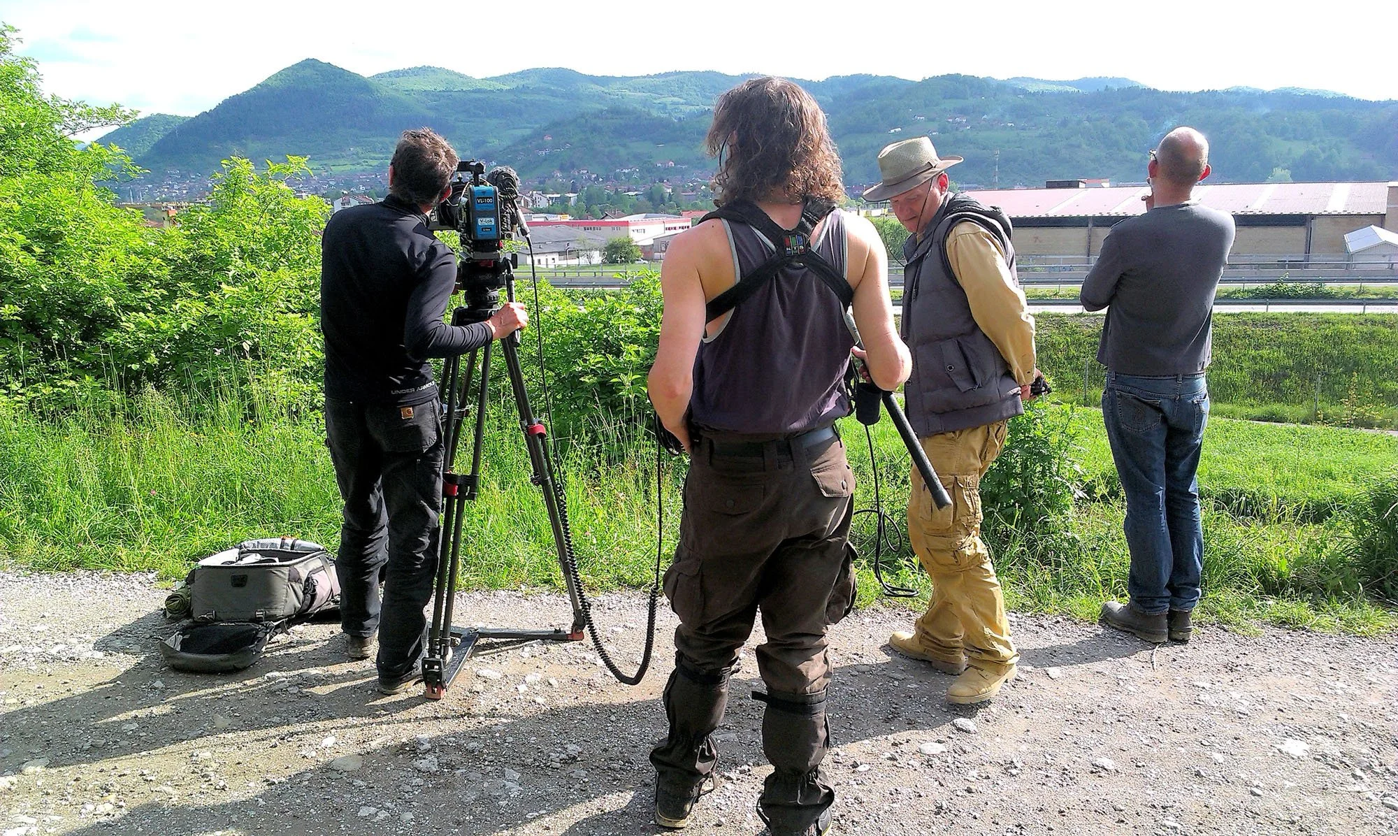 A group of five people, including a woman and four men, are standing outdoors on a dirt and gravel path, with a green hillside and industrial buildings in the background. One person is operating a professional video camera mounted on a tripod, while 