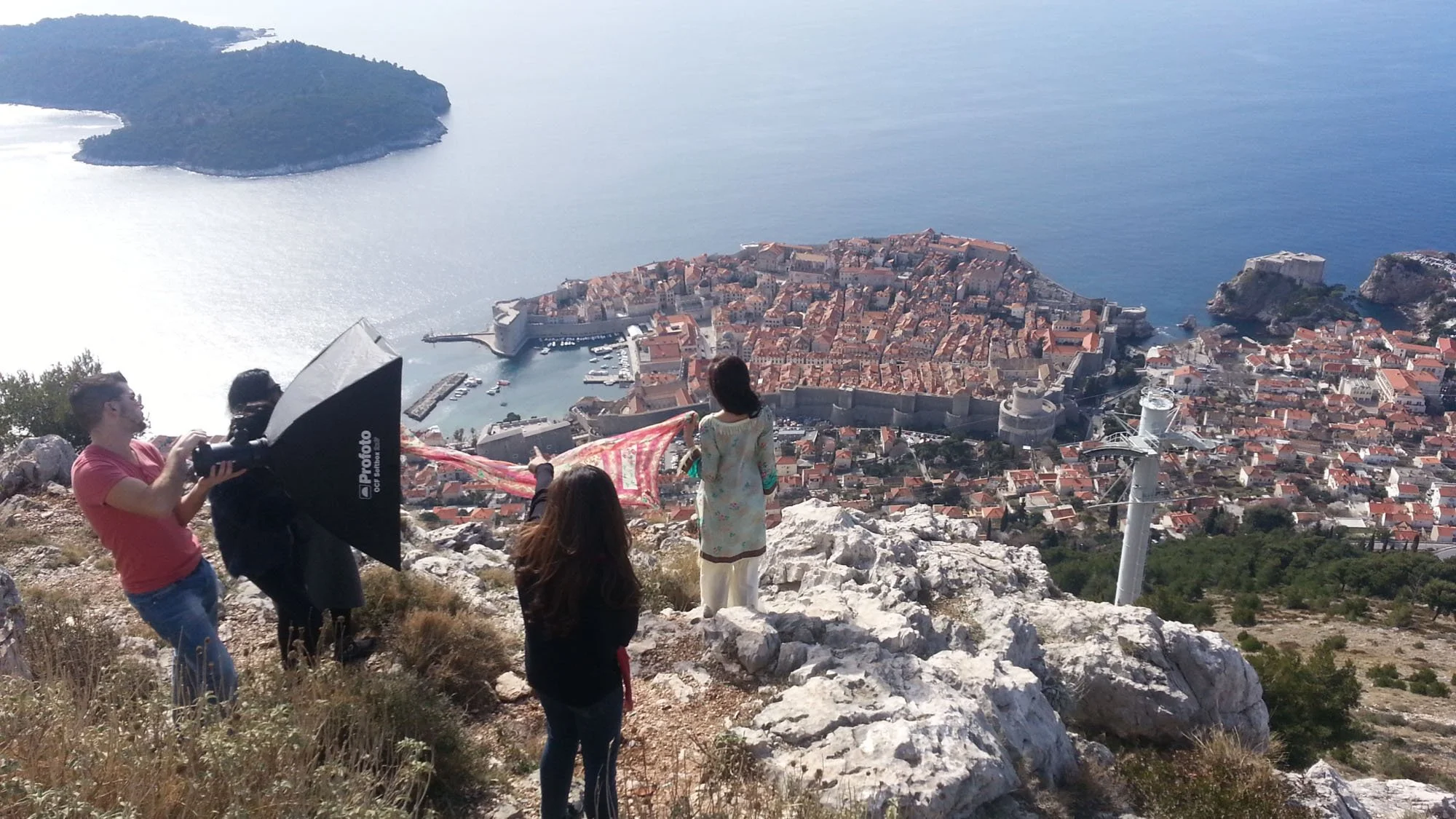 Photographers take pictures of a historic town with fortified walls and red-tiled roofs from a mountain viewpoint overlooking the Adriatic Sea.