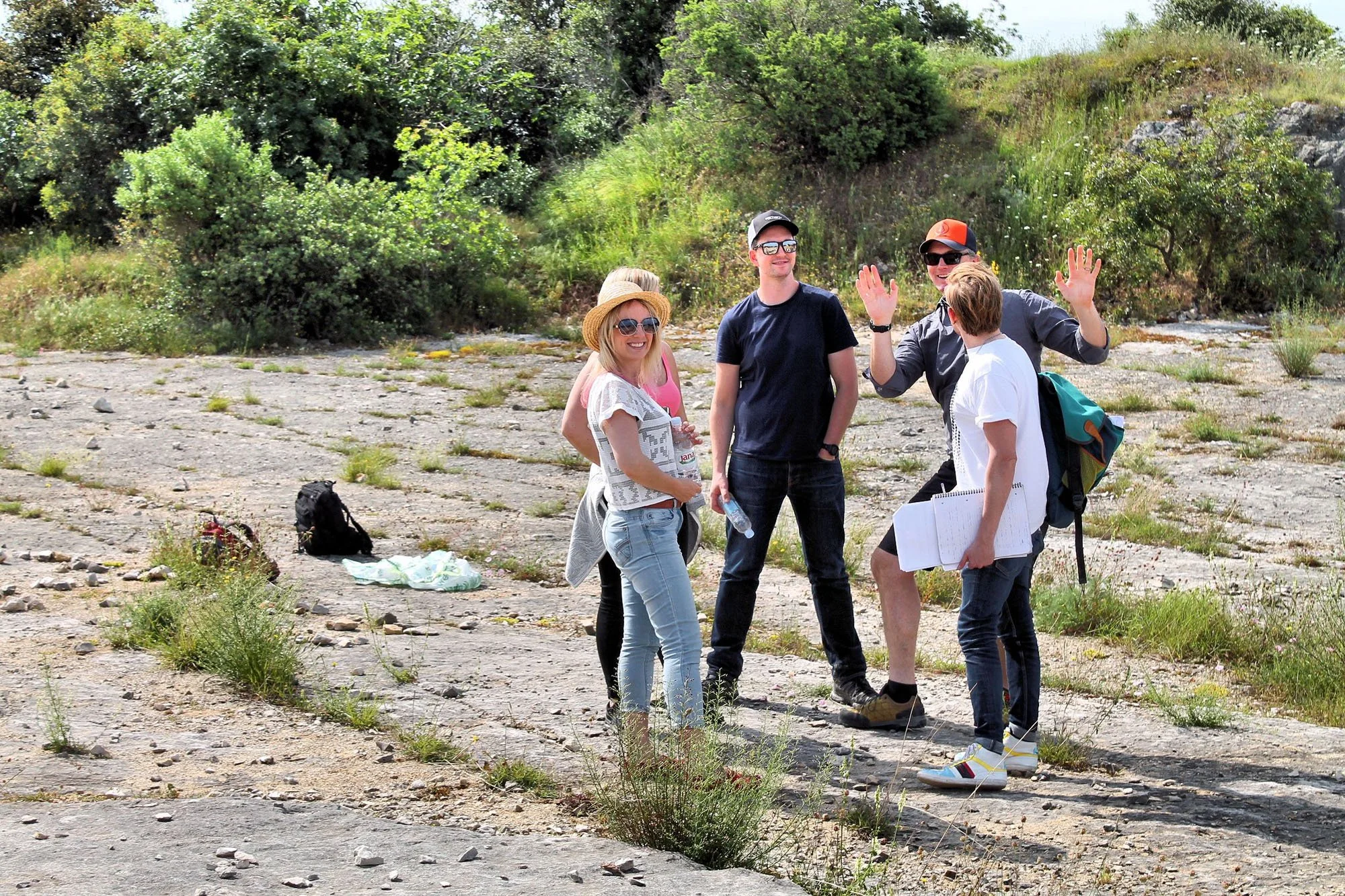 Five people standing and talking outdoors, some wearing sunglasses and hats, amid a natural landscape with gravel, grass, and green bushes.