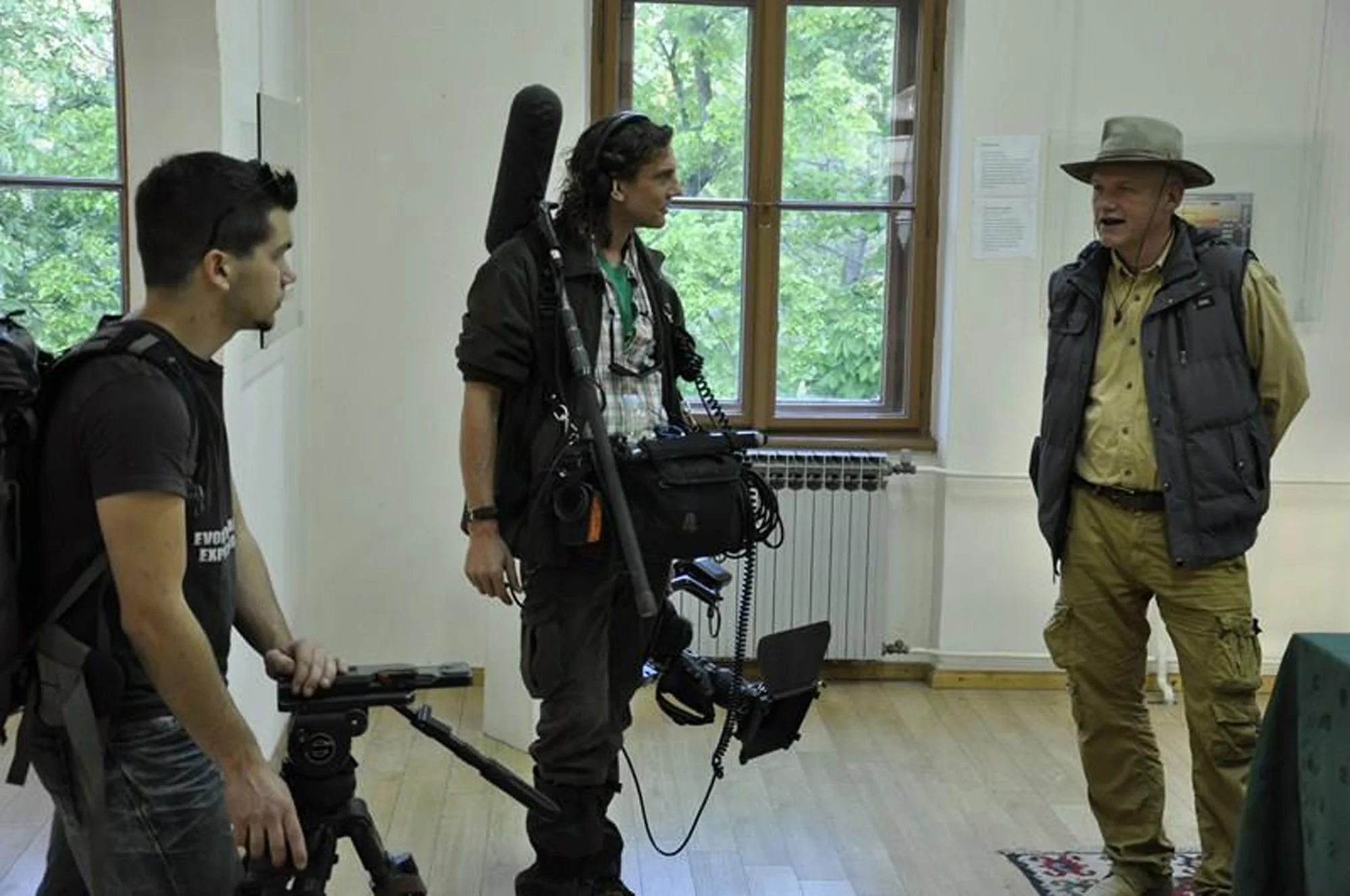 Three people are standing in a room with white walls and large windows. The person on the left is a young man with dark hair, wearing a black shirt and carrying a backpack. The person in the middle is a woman with curly hair, wearing outdoor gear inc