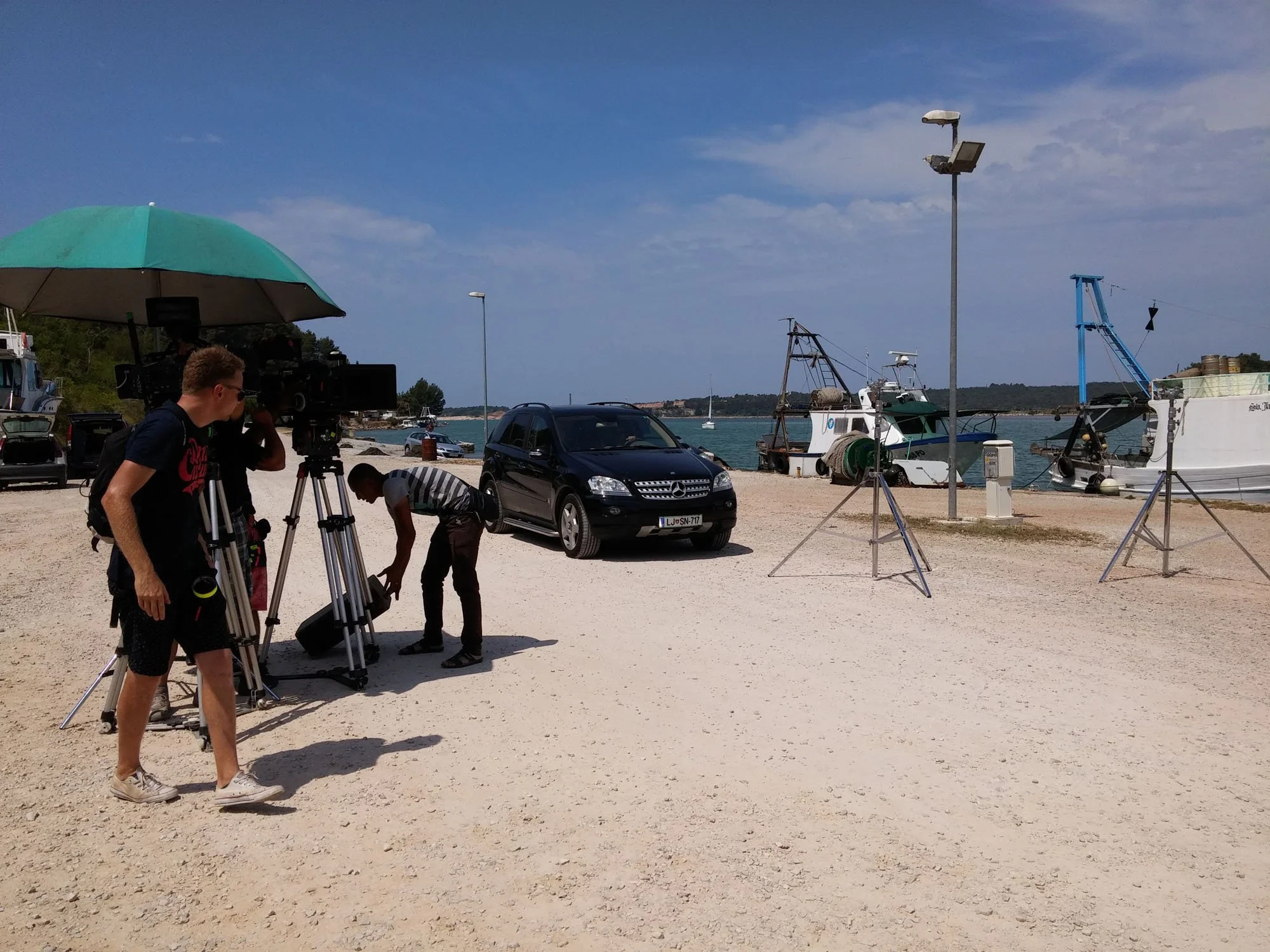 A team of filmmakers setting up equipment on a beach by the water, with boats docked nearby and a clear blue sky overhead.