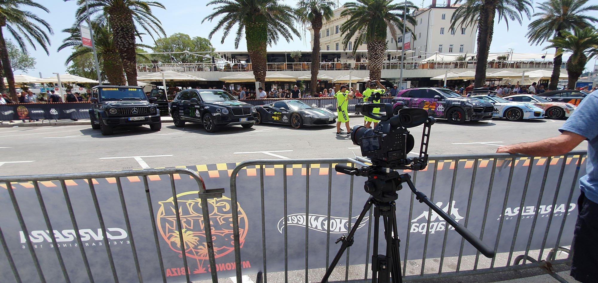 A row of luxury and sports cars parked on a street during a car event, with people walking and filming. Palm trees and a building are in the background, and there is a camera on a tripod in the foreground.