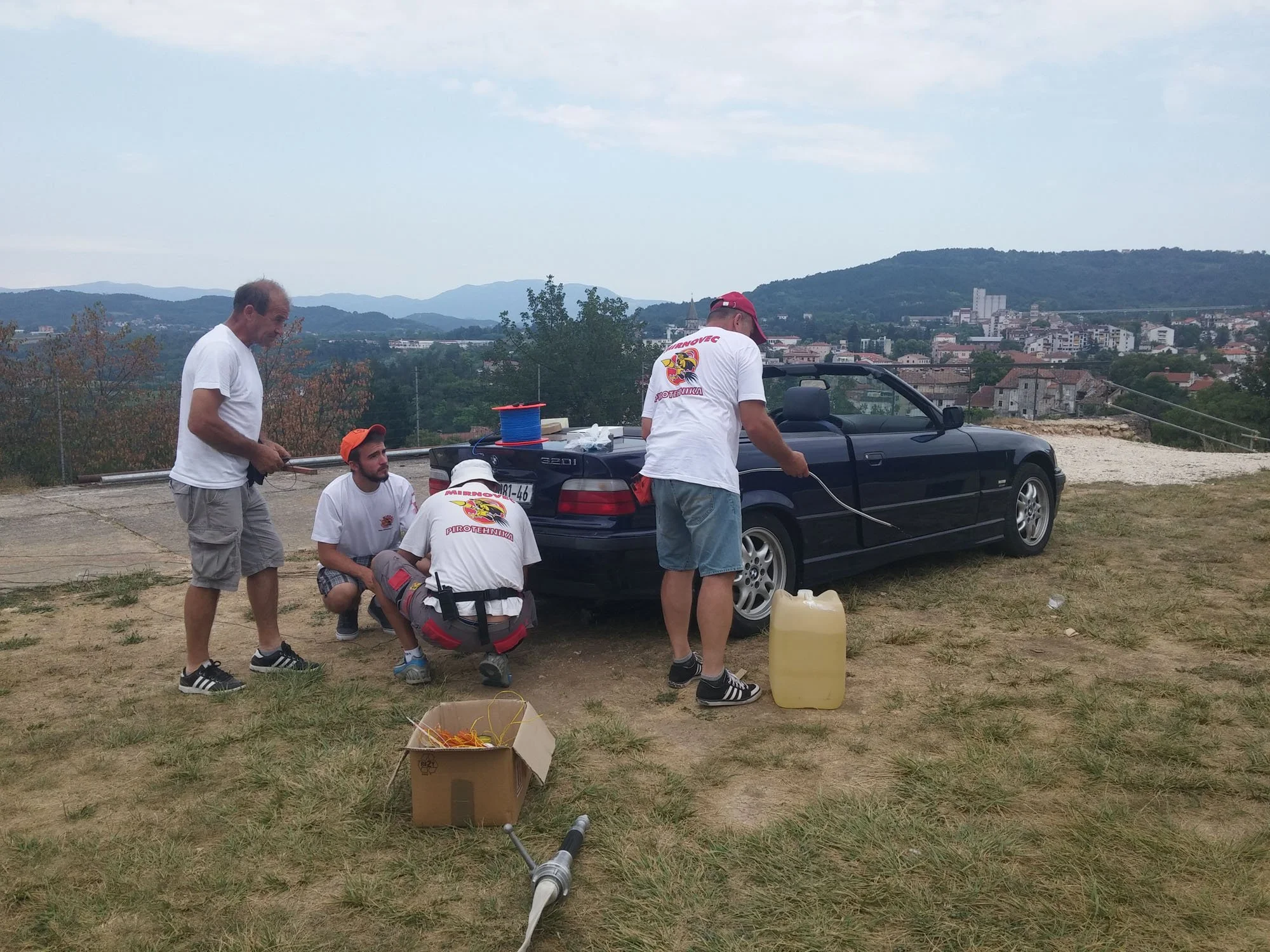 A group of five men working outdoors on equipment near a black car with an open trunk, surrounded by a grassy area with mountains and a town in the background.