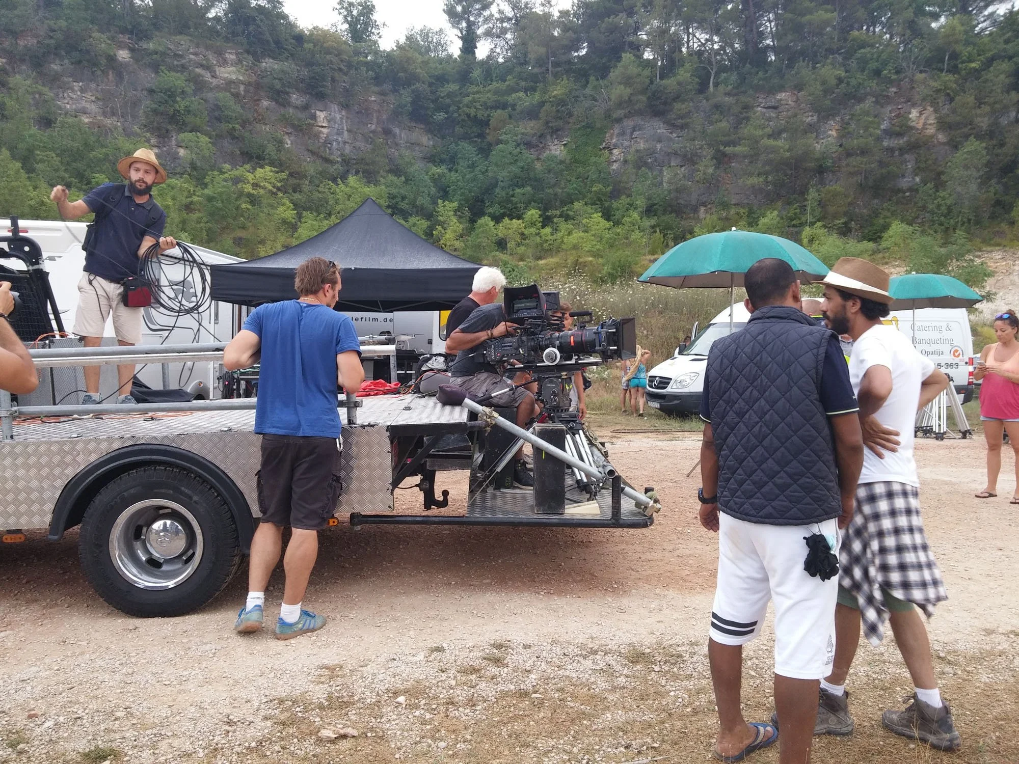 Filming crew setting up equipment outdoors in a mountainous area with trees and cliffs in the background, multiple people preparing for a shoot.