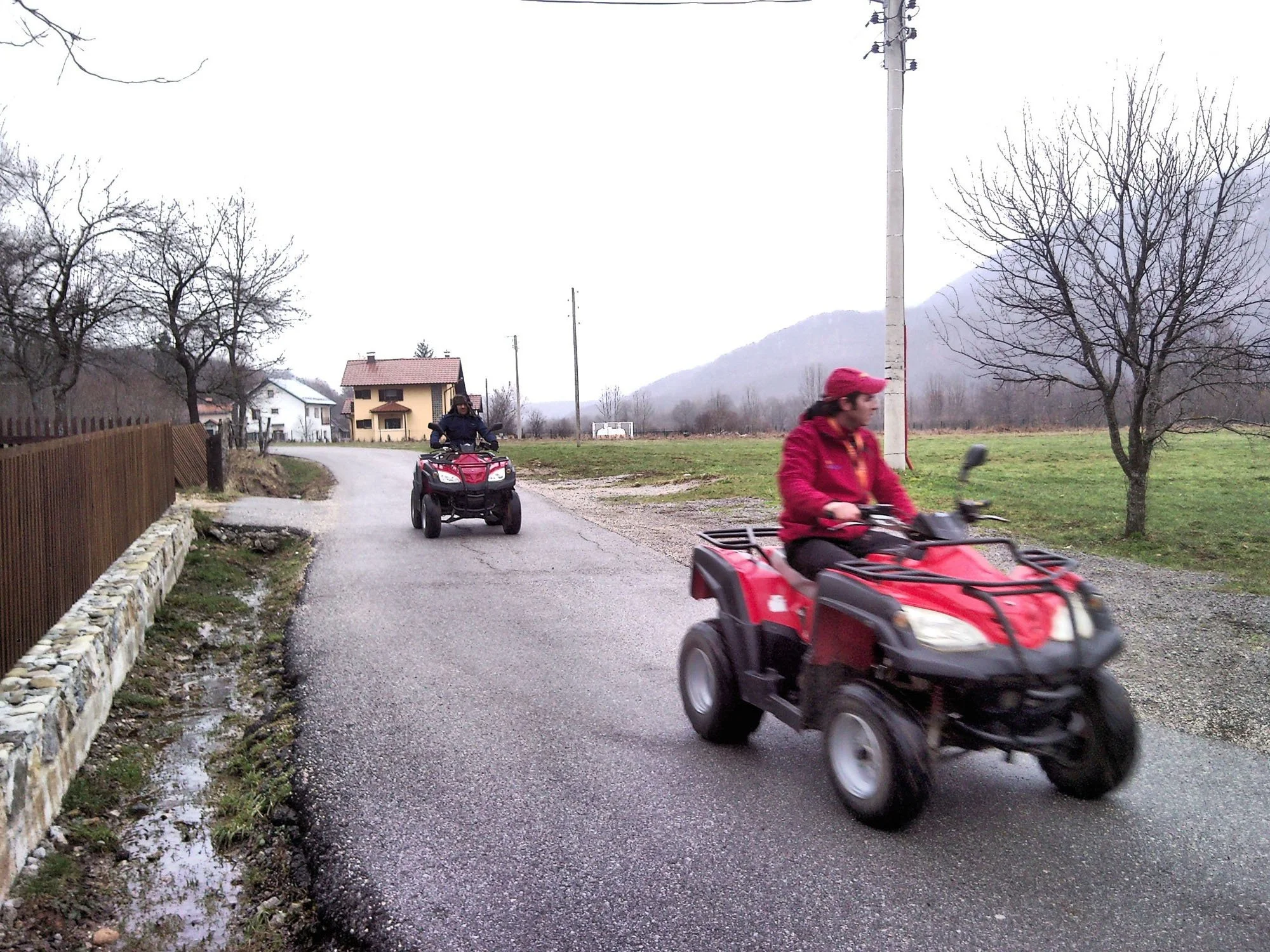 Two people riding all-terrain vehicles (ATVs) on a rural road with houses and leafless trees, under an overcast sky, in a mountainous area.