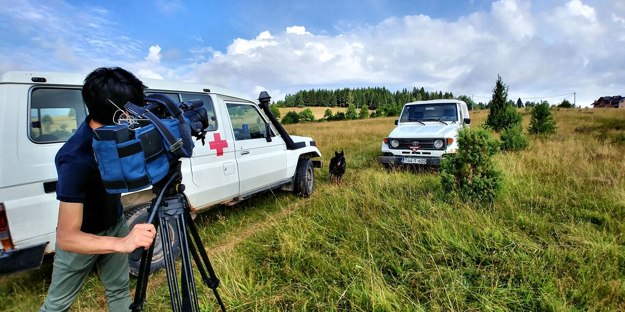 A person operating a video camera in a grassy field with two parked vehicles, an ambulance and a white truck, and a black dog between them, with trees and a house in the background under a partly cloudy sky.