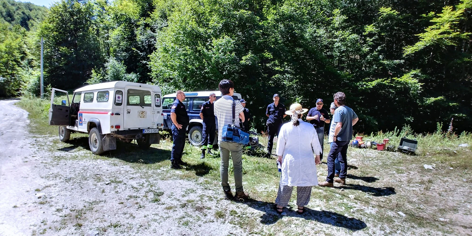 Group of people, including emergency responders and civilians, gathered outdoors on a grassy and gravel area near vehicles under green trees, with equipment and supplies nearby.