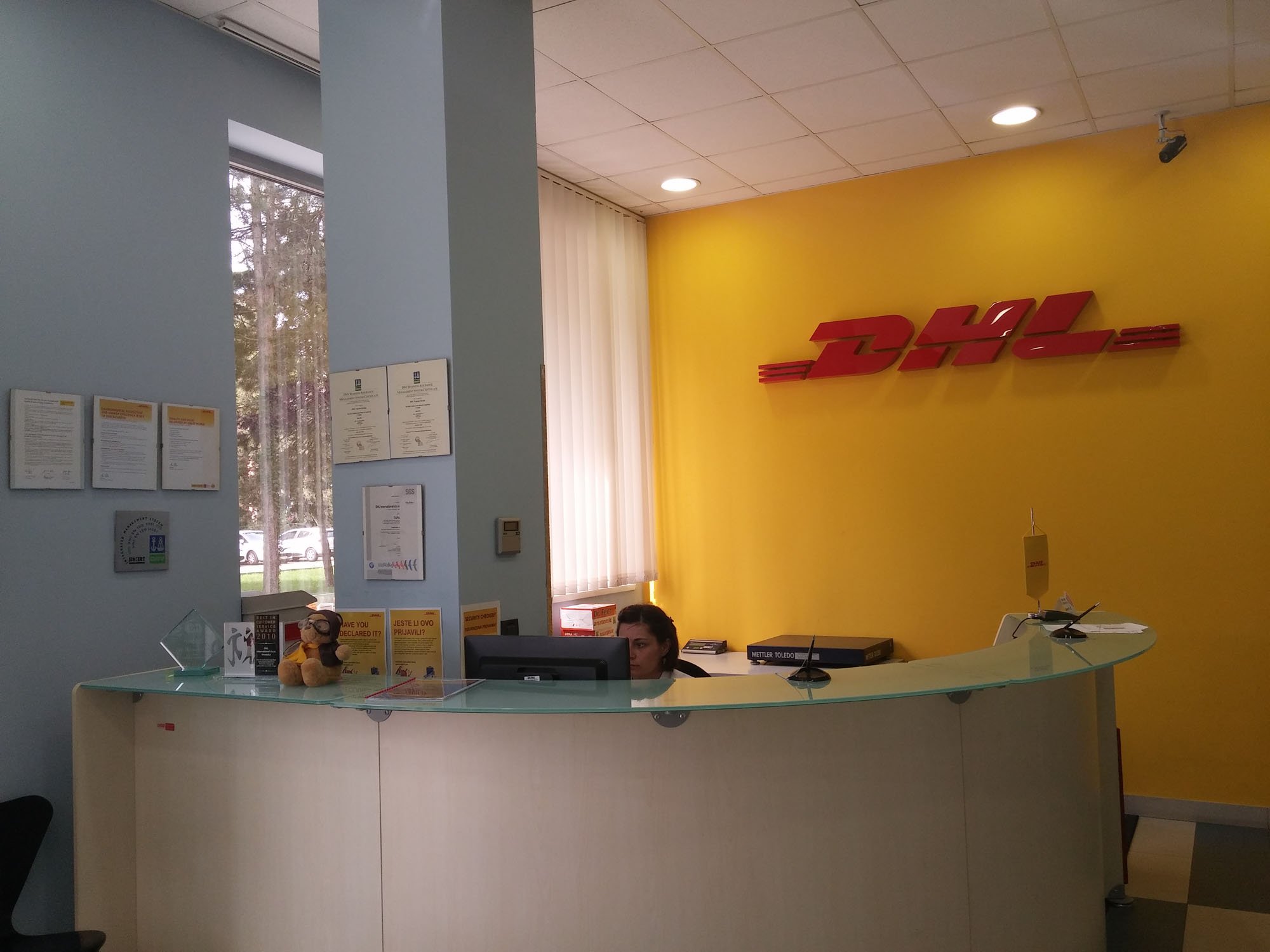 DHL office reception desk with a yellow wall behind it displaying the red DHL logo and a window with vertical blinds. A woman is sitting at the desk, and another person is visible in the lower-left corner.
