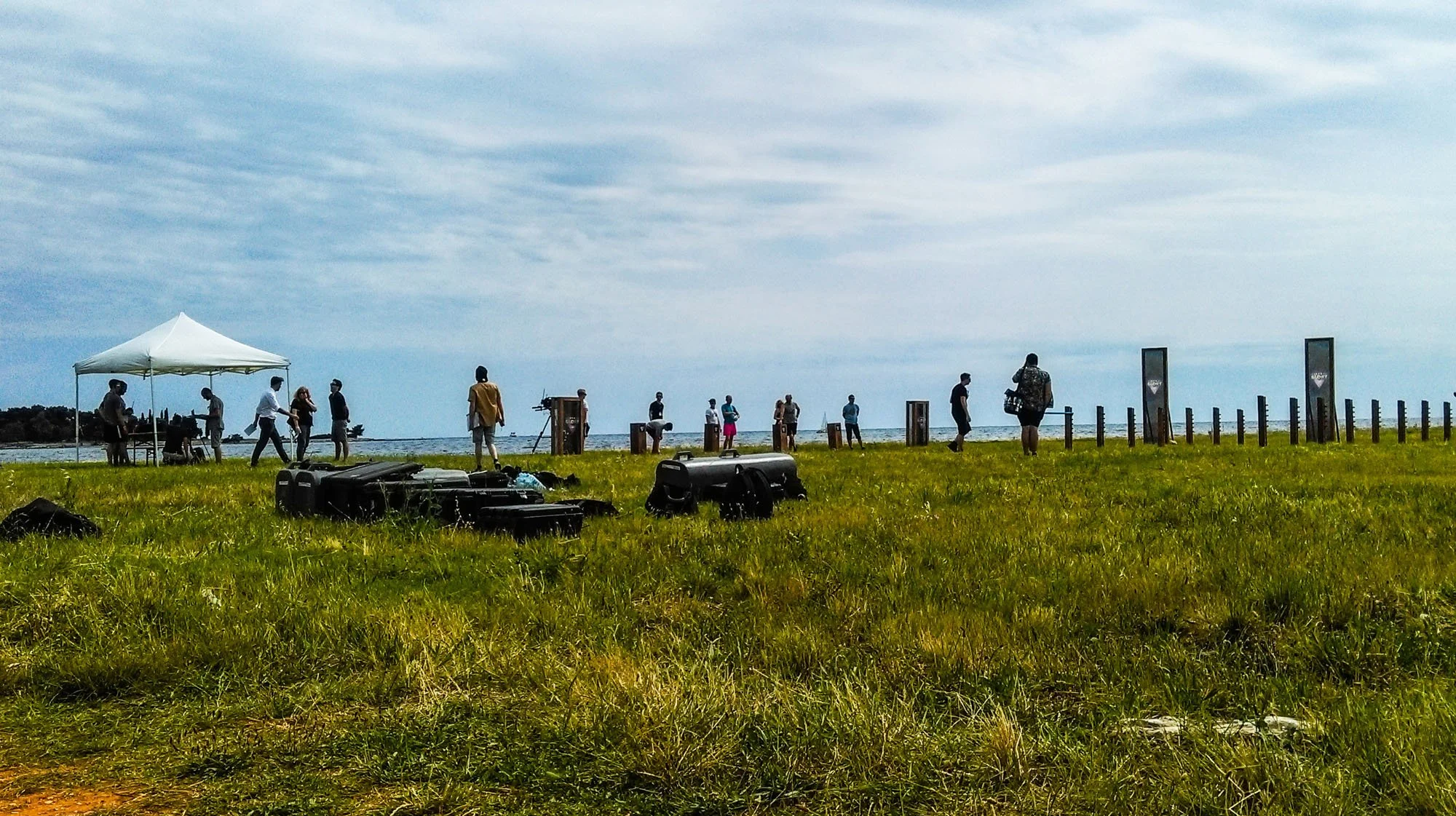 A group of people gathered on a grassy field near a body of water, with equipment and a white canopy tent, under a partly cloudy sky.
