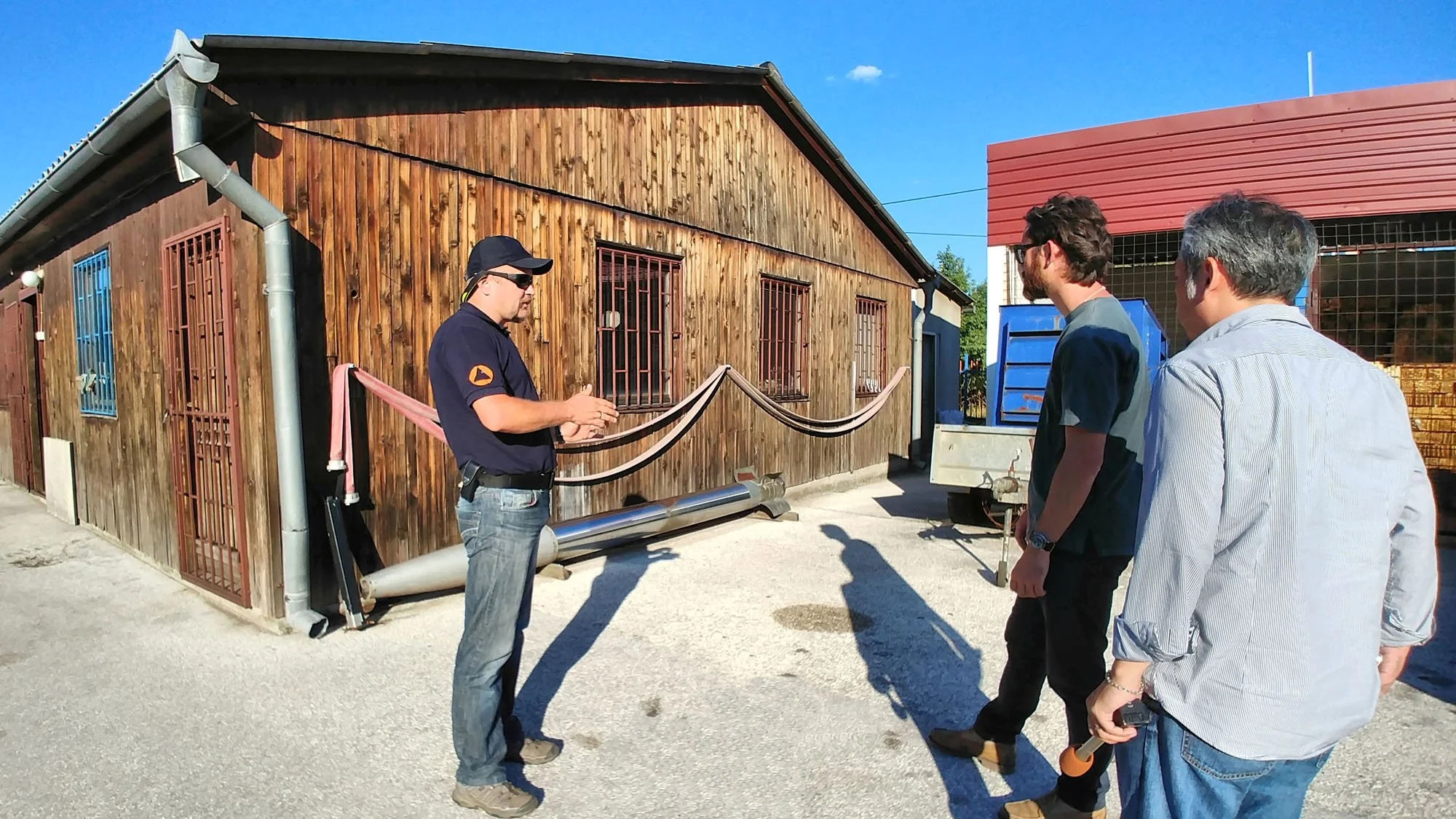 A group of three men standing outdoors in front of a wooden building during daytime, with a man in uniform talking to them. The scene appears to be a guided tour or educational activity.