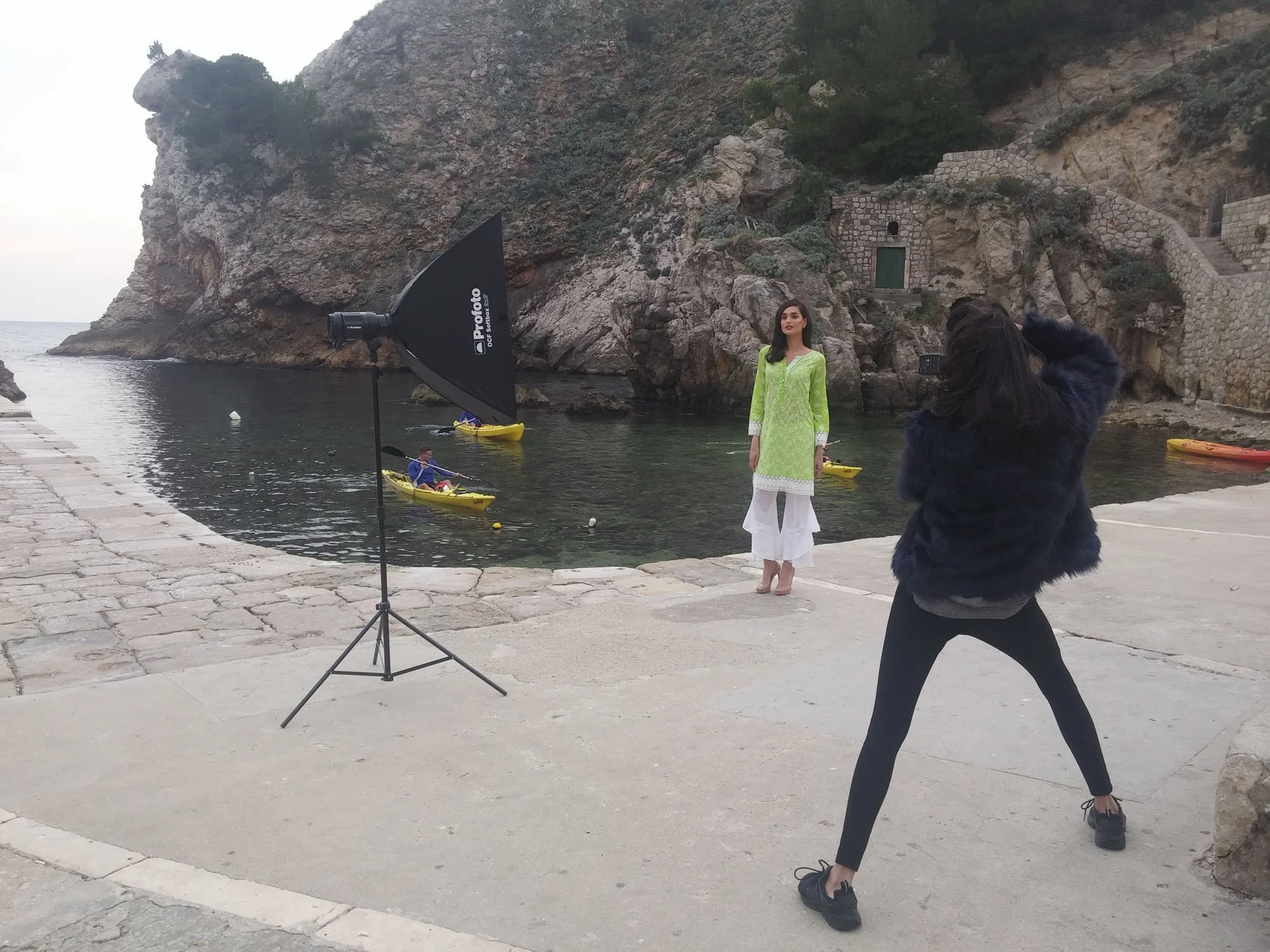 A photo shoot near a rocky shoreline with a woman in a green traditional dress posing for photos, a photographer taking her picture, and a large softbox lighting the scene. There are kayakers in the water and rugged cliffs in the background.