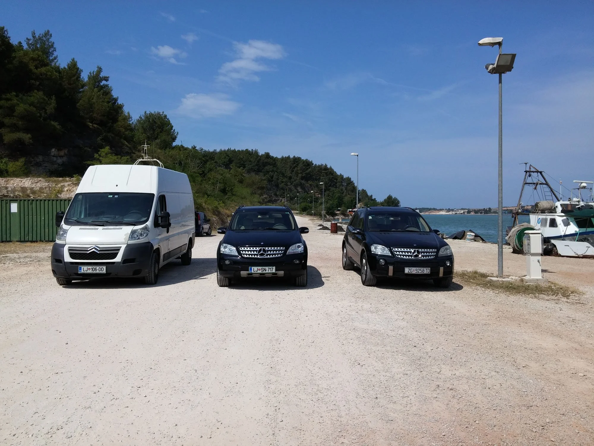 View of a parking area near a body of water with several parked cars, including a white van and black vehicles, surrounded by a green fence, streetlights, boats, and a rocky hillside with trees under a partly cloudy blue sky.