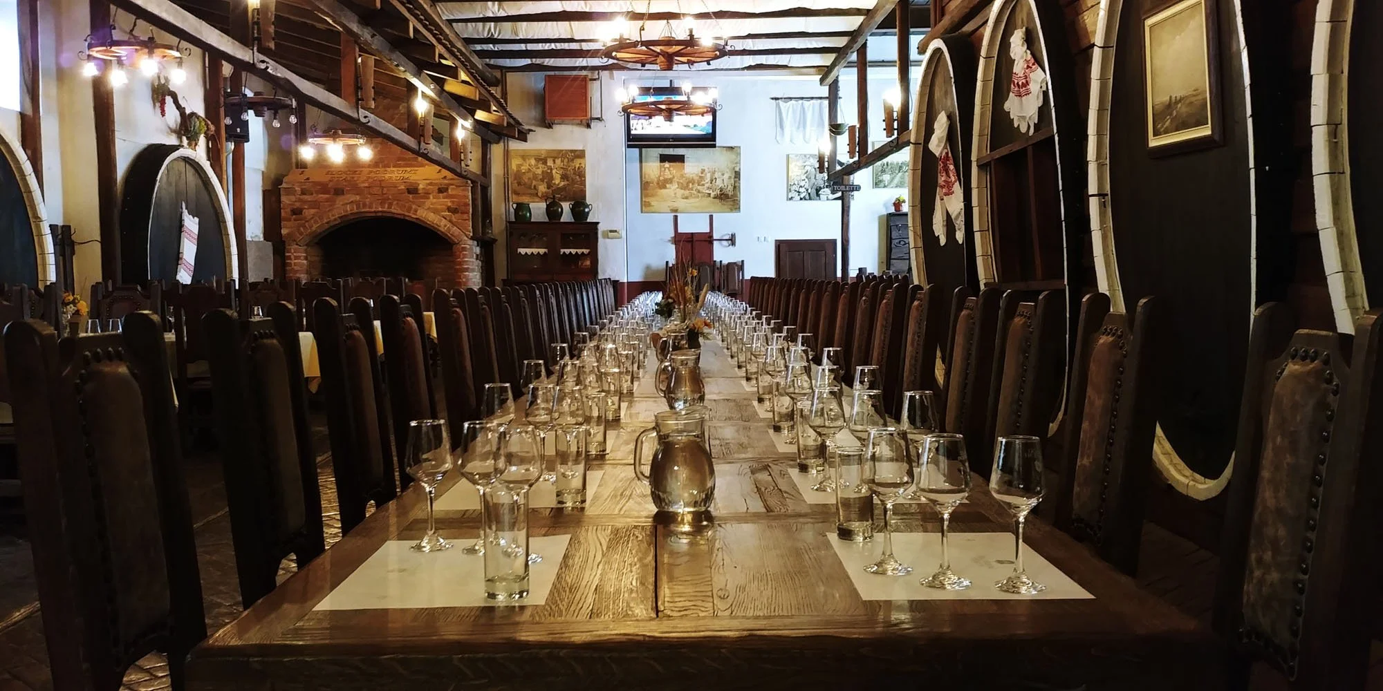 Long wooden dining table set with wine glasses, water jugs, and floral centerpieces in a rustic, cozy restaurant with wooden chairs and brick fireplace.