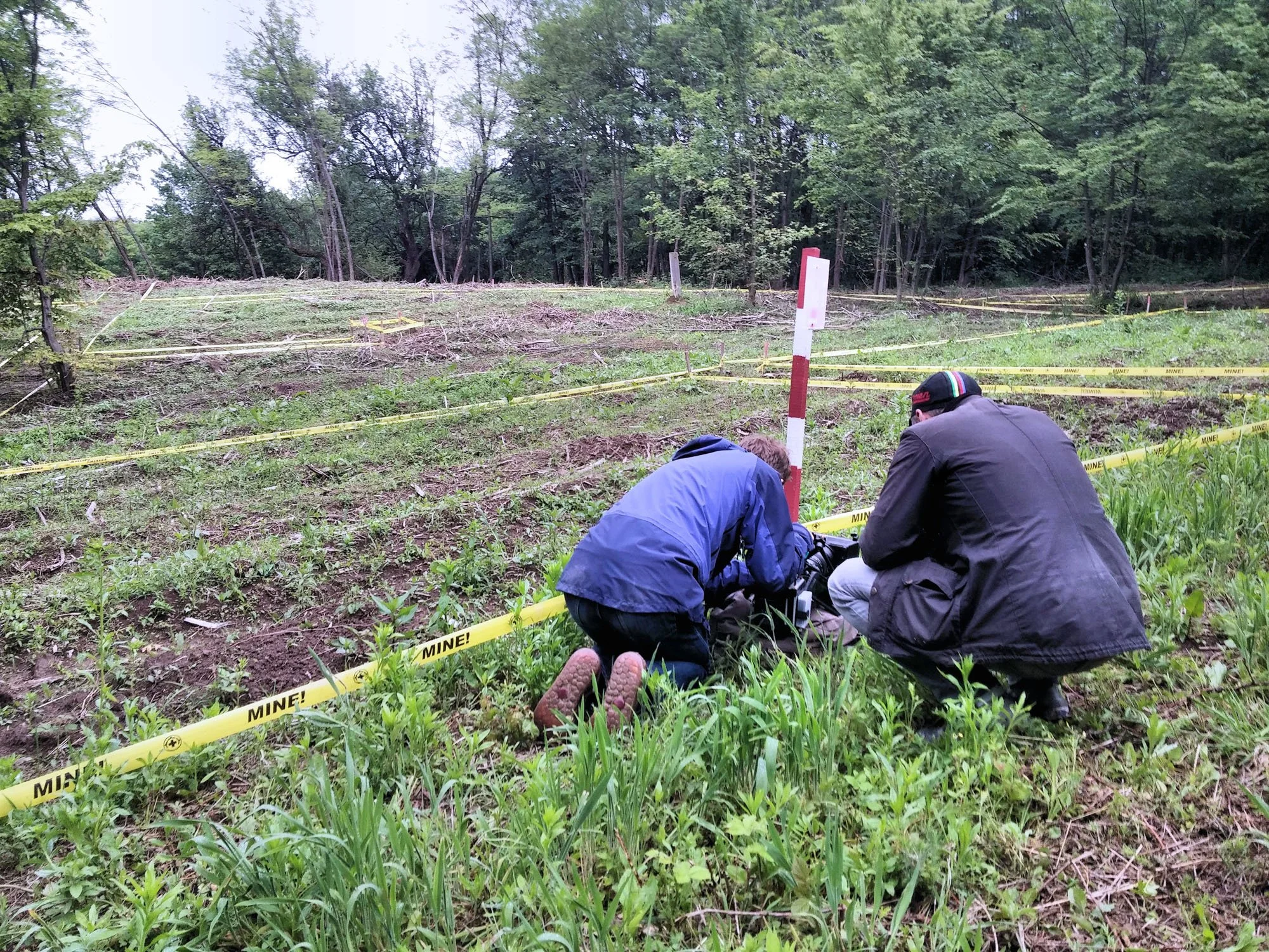 Two men working at soil excavation site surrounded by yellow 'MINES!' tape with a forest background.