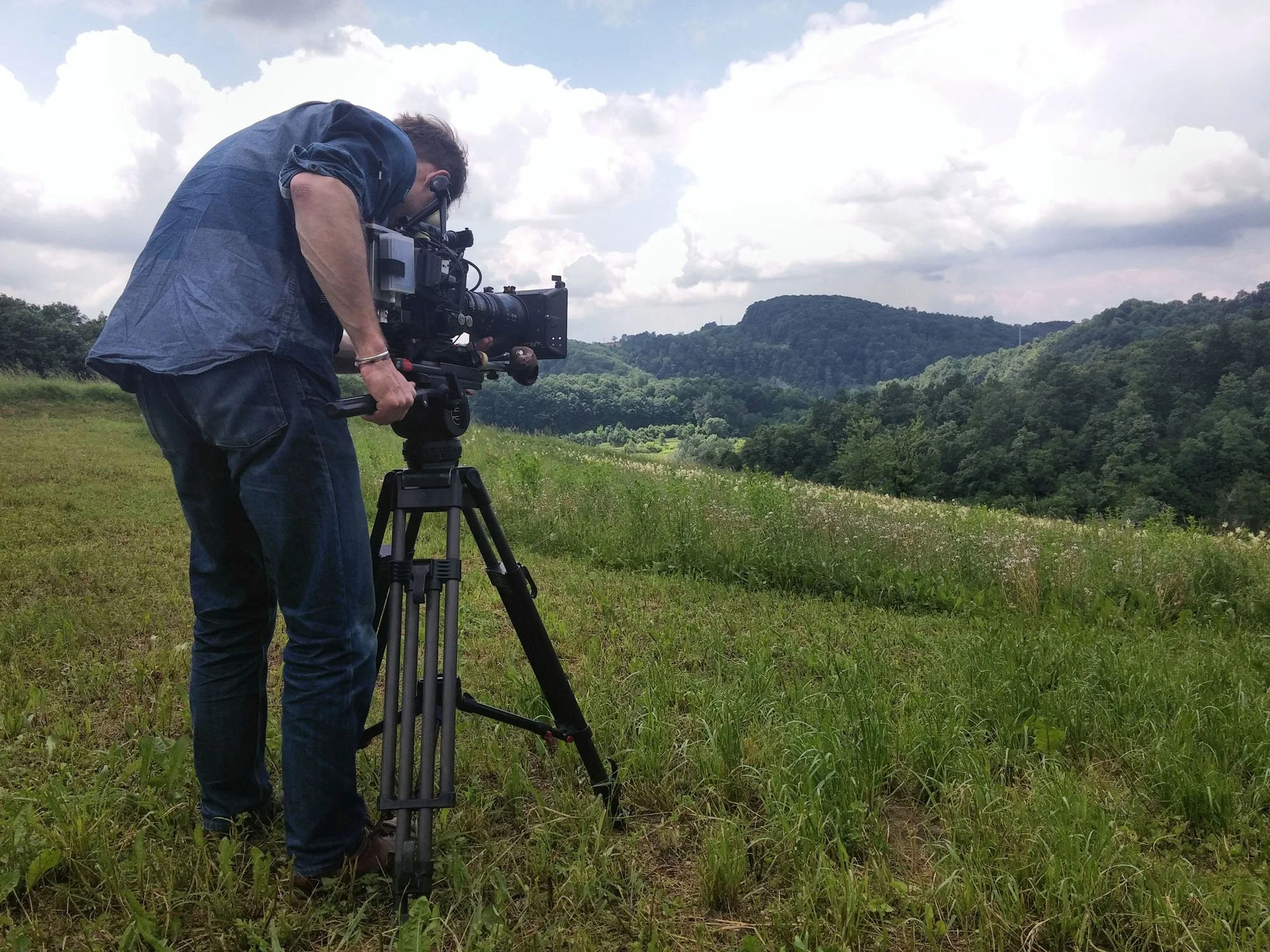 A man operating a professional video camera on a tripod outdoors in a green, hilly landscape under cloudy skies.