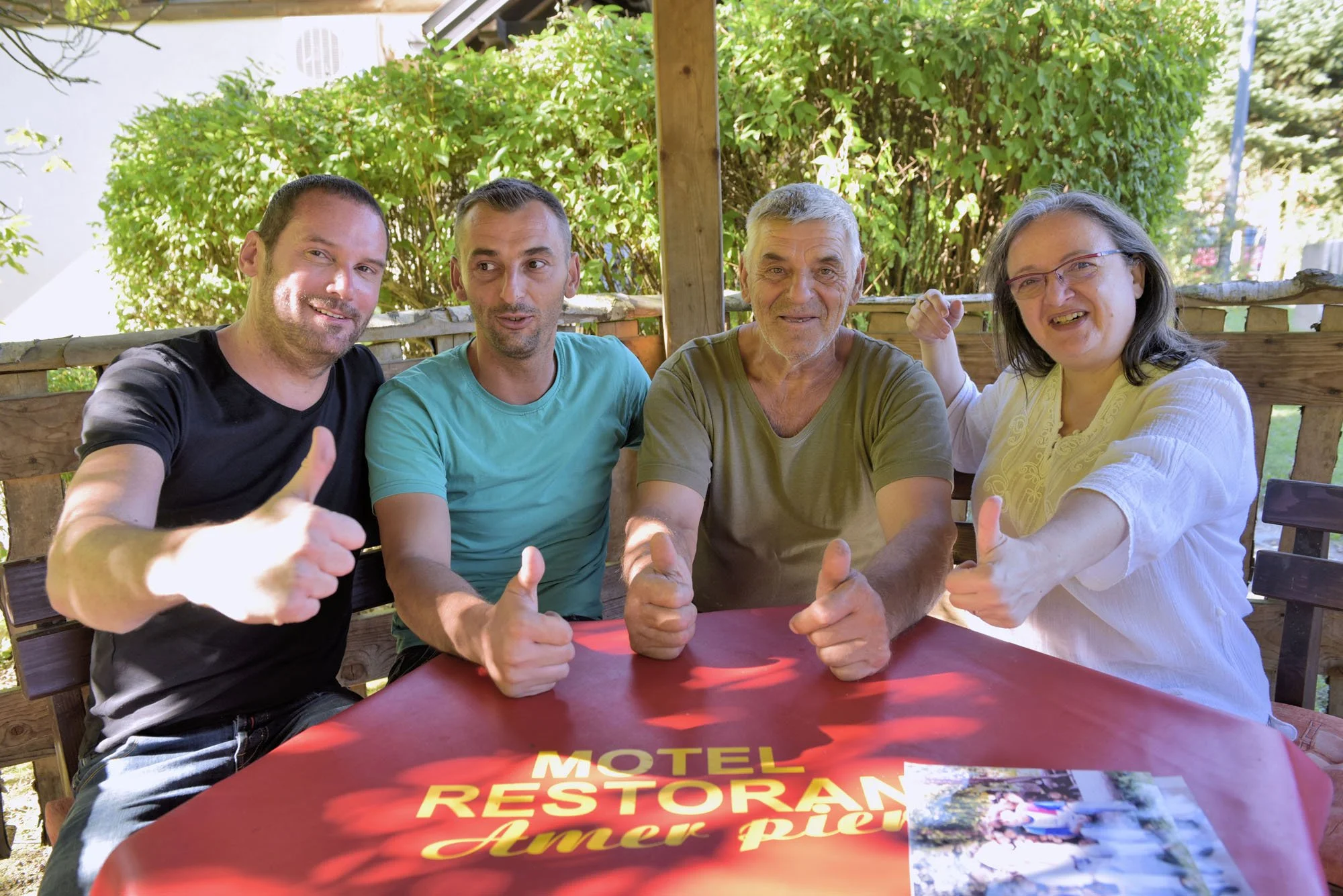 Four people sitting at a table outdoors, smiling, giving thumbs up, with a lush green background. The table has a red tablecloth with yellow text, partially visible, reading "MOTEL RESTORAN..." and a photo of a scenic landscape.
