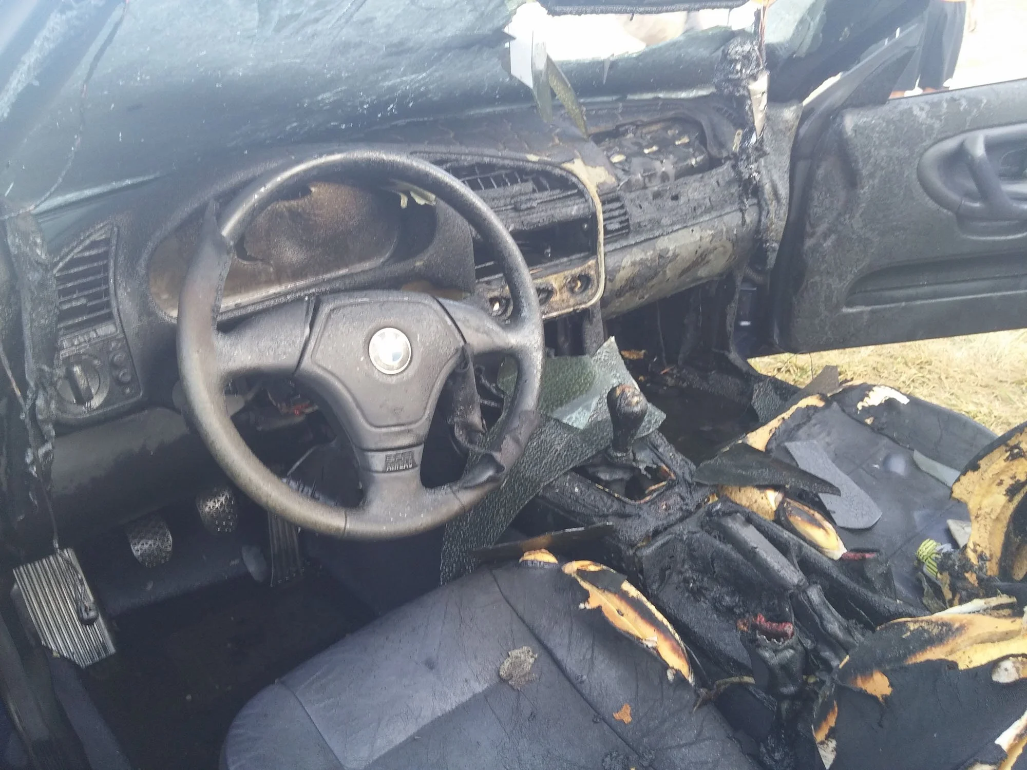 The interior of a car shows severe fire damage. The dashboard, steering wheel, and seats are charred and melted. The car appears to have been in a fire, with burning and soot covering the surfaces.