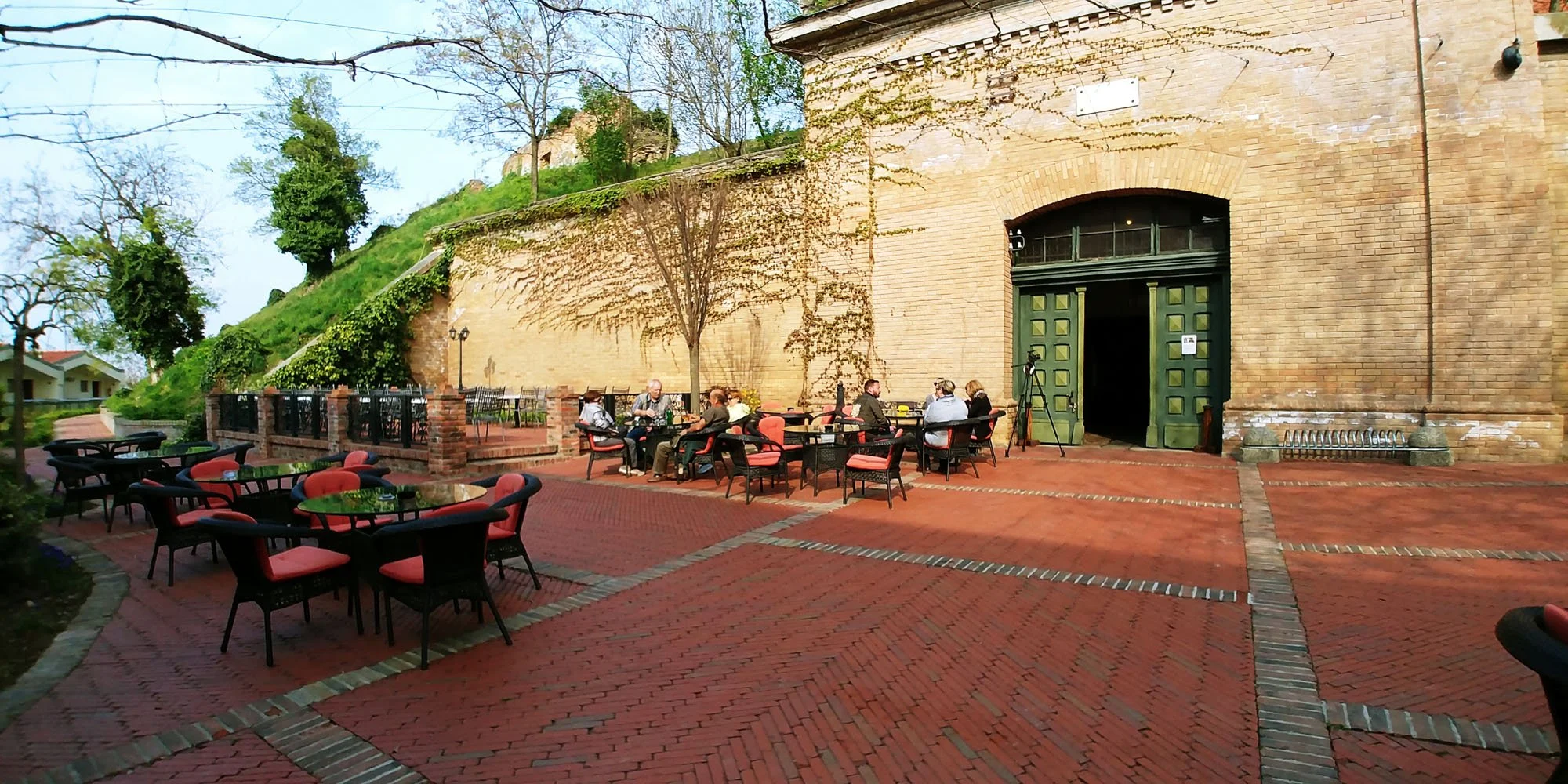 People sitting at tables outside a historic brick building with large open doors, surrounded by greenery and a hillside.