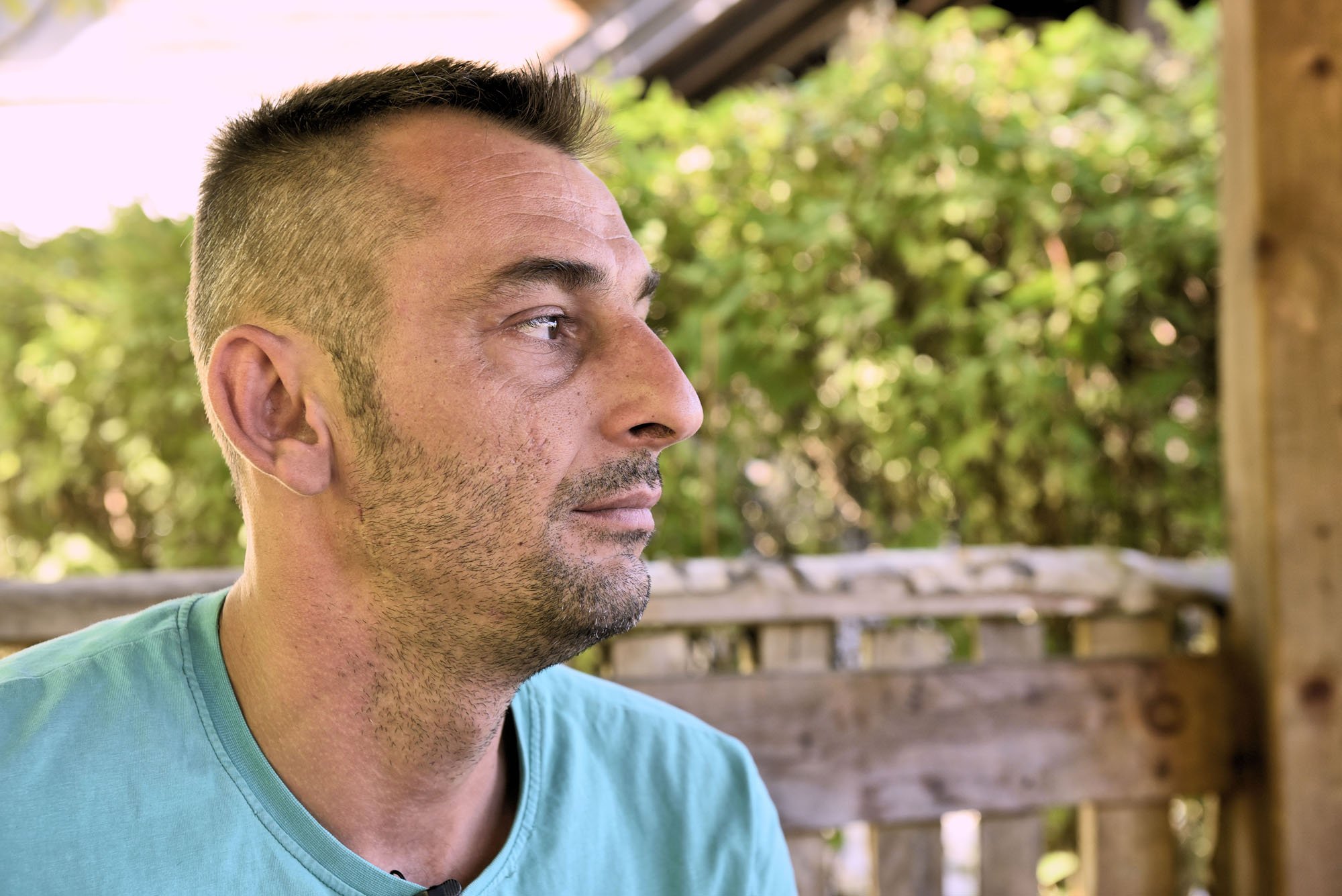 Profile view of a middle-aged man with short dark hair, a goatee, and wearing a teal T-shirt, sitting outdoors with green foliage and a wooden fence in the background.