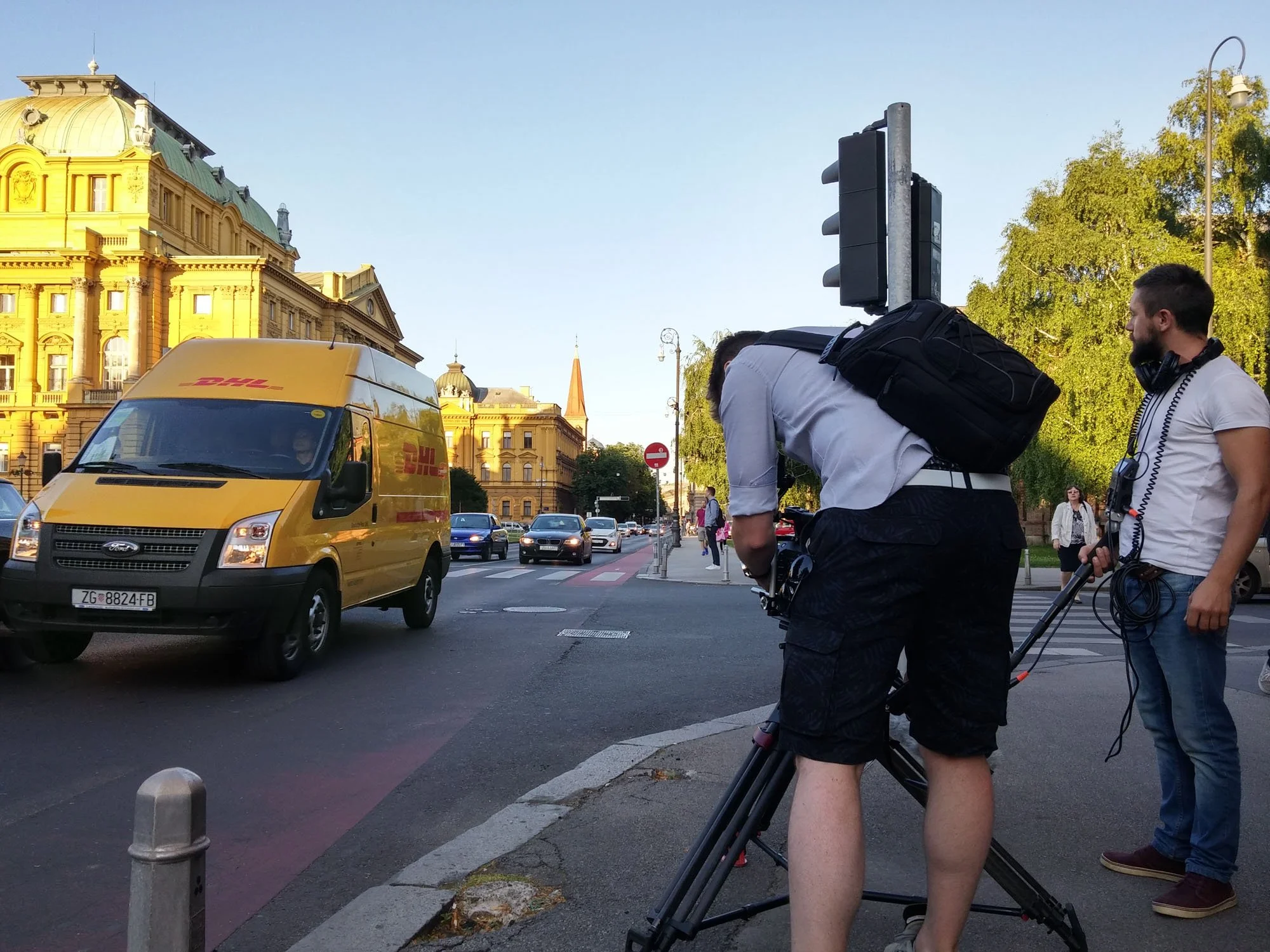 Street scene with a yellow DHL delivery van, people working with filming equipment, including a person adjusting a camera on a tripod, and a man with headphones and a backpack, in front of a historic yellow building with a green roof and a blue sky.