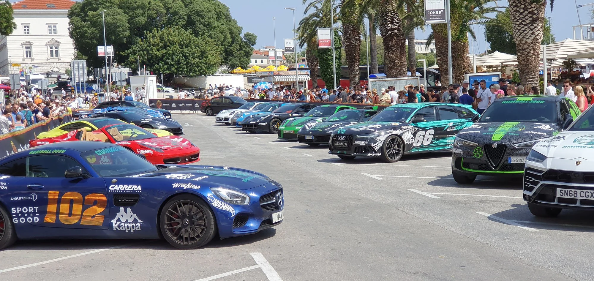 Various luxury and sports cars parked in a row at a crowded outdoor event with people and palm trees in the background.