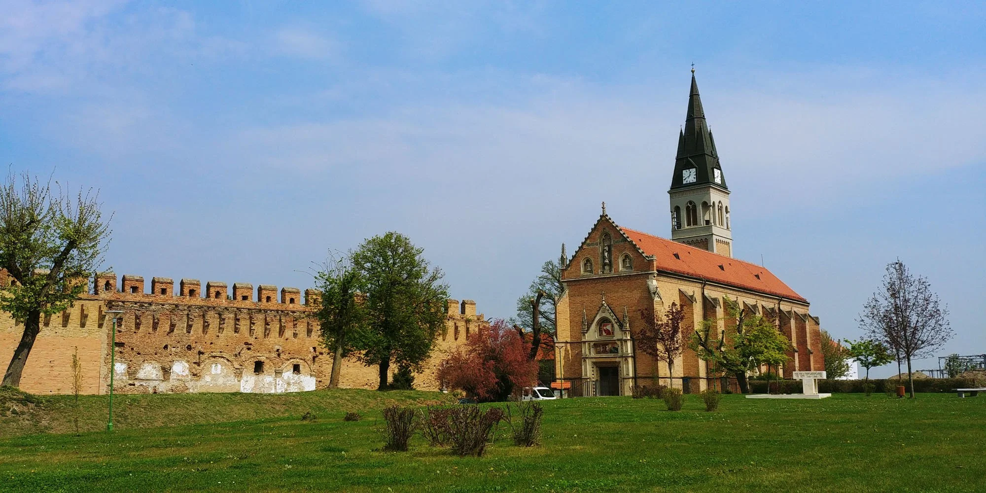 A church with a tall steeple and a red roof, surrounded by a grassy area with trees and a brick wall in the background, under a partly cloudy sky.