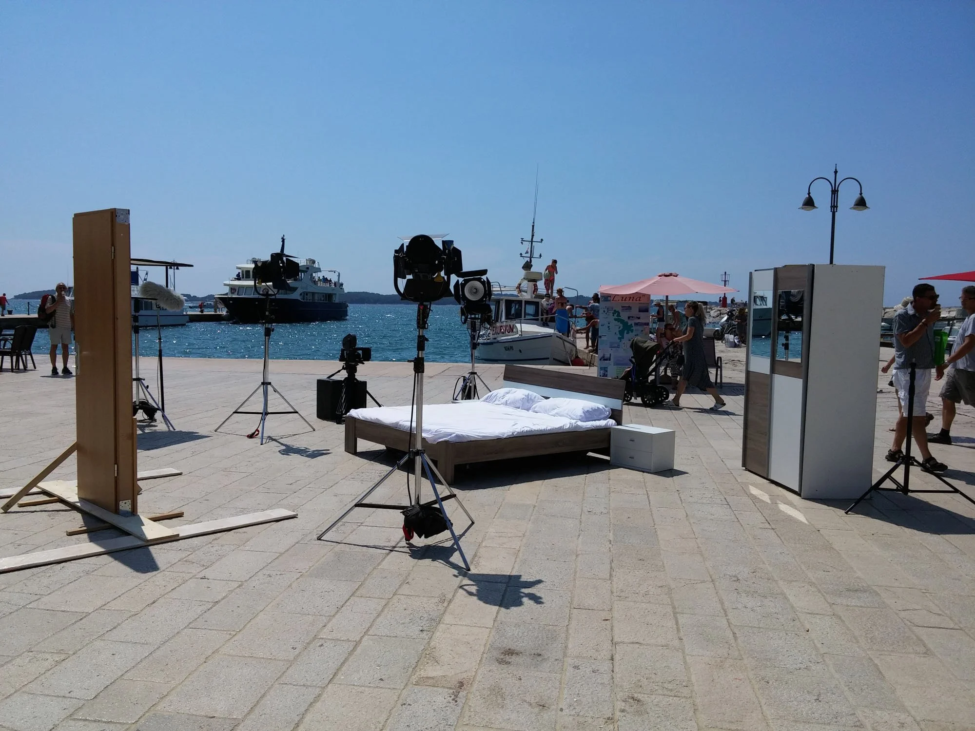 A sunny beachside scene with a viewing setup, featuring a bed and photo equipment in the foreground, boats on the water, and people walking on the coast under clear blue skies.