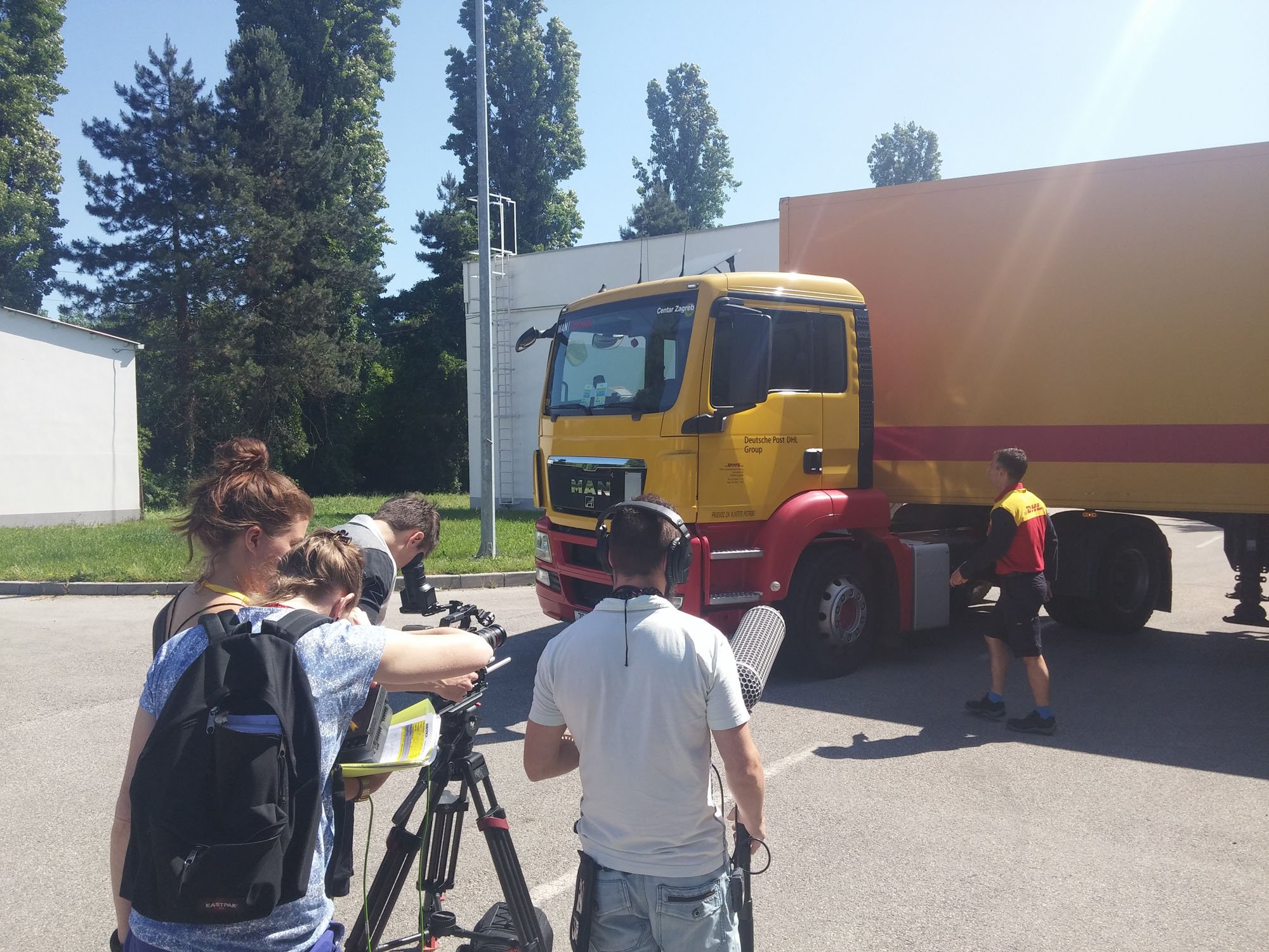 A group of people, including a person with headphones and camera equipment, appear to be filming or photographing a yellow and red delivery truck parked outdoors on a sunny day. Several large trees are visible in the background.