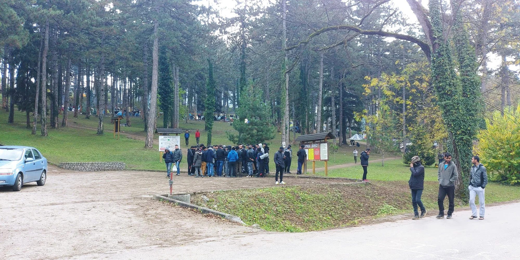 People gather in a park surrounded by tall trees, some waiting in line, others walking, and a few taking photos on a cloudy day.