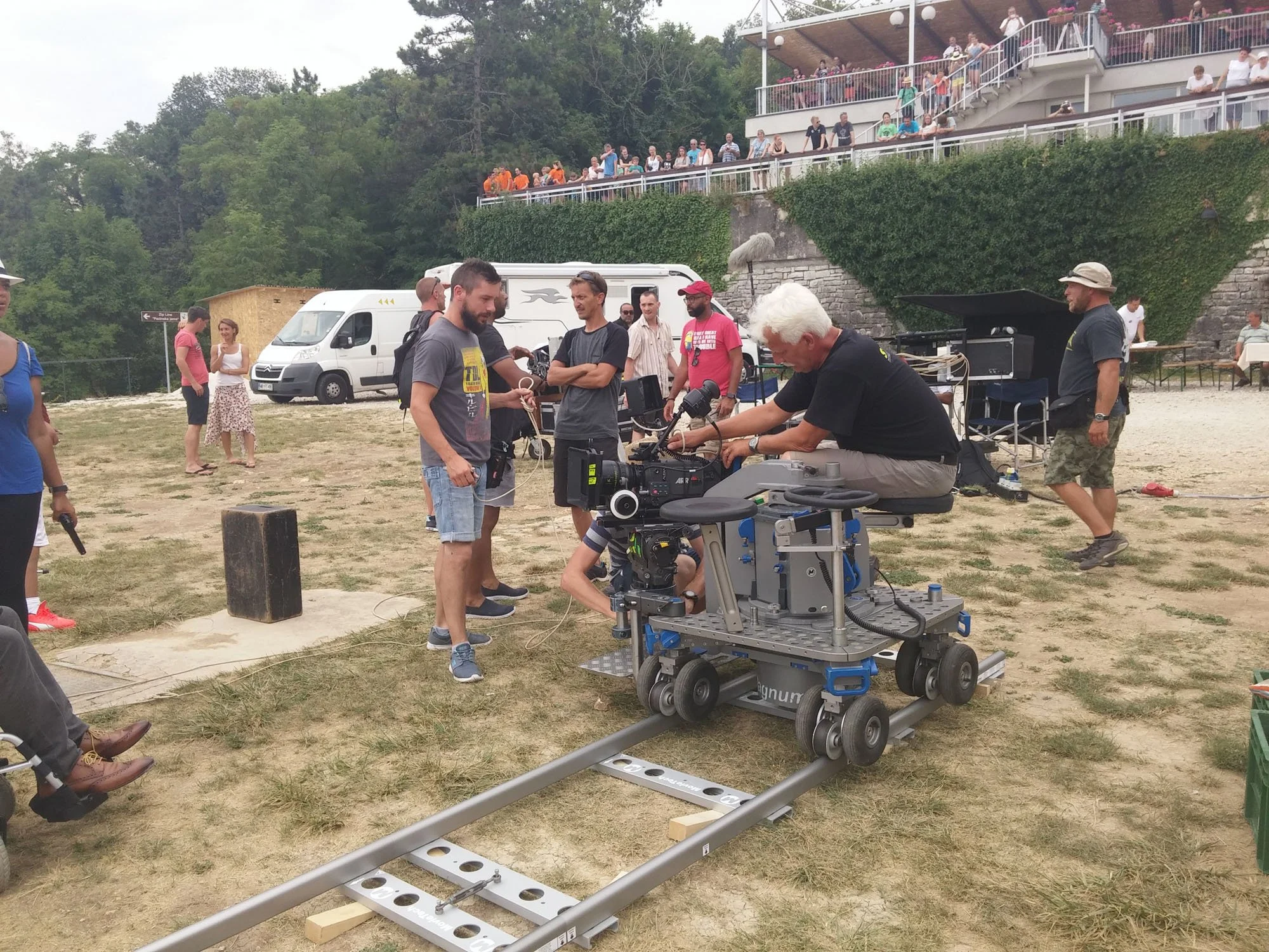 People on a film set working with a camera on a dolly outdoors, with an audience watching from a terrace above.