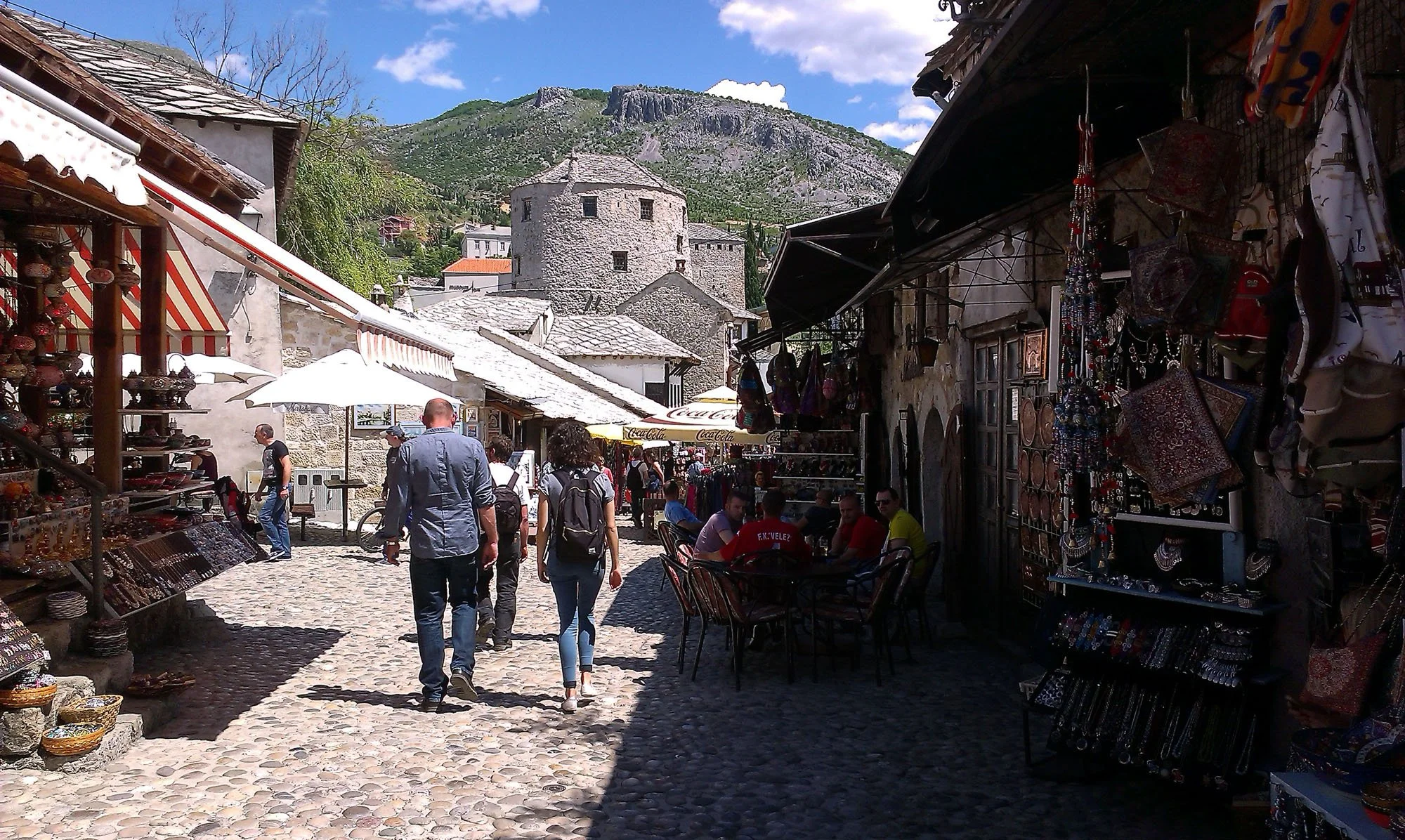 A busy outdoor marketplace with vendors selling souvenirs and crafts, with people walking and sitting at tables, against a backdrop of historic stone buildings and a mountainous landscape.