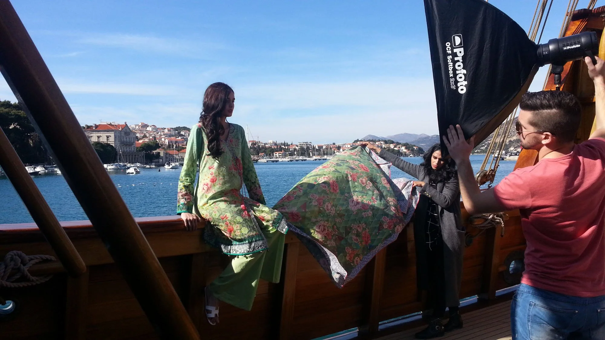 A photo of a woman in a floral dress on a boat, holding an umbrella, with a crew member adjusting lighting equipment, and a scenic harbor with boats and mountains in the background.