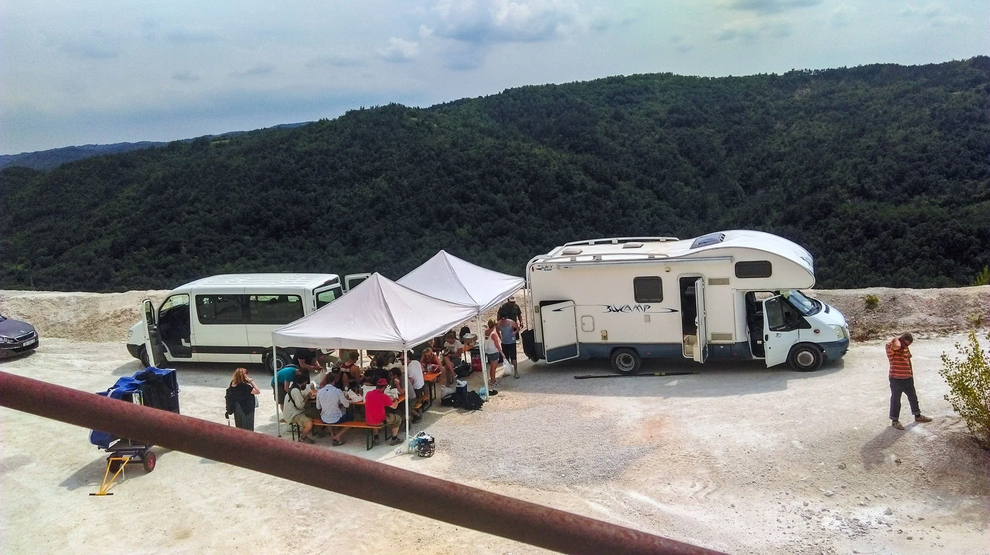 A group of people gathered under a white canopy tent, sitting at picnic tables, near parked camper vans and cars, with green hills and a cloudy sky in the background.