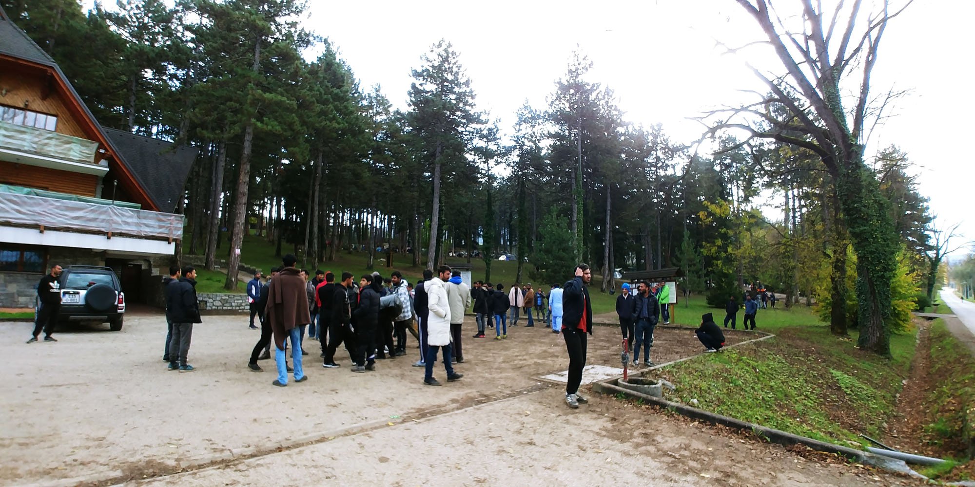 A group of people gathered outdoors near a wooded area with tall trees, some in jackets and casual clothing. There is a building on the left with balconies and a parked car nearby. The scene appears to be in fall, with some people engaged in conversa