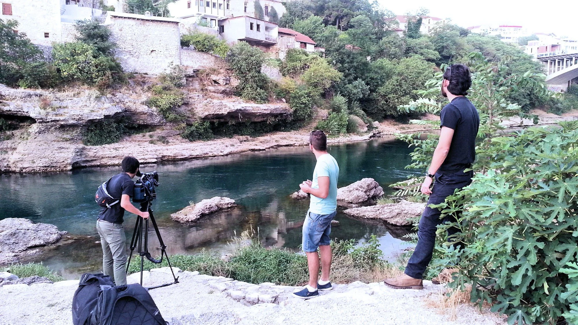Three men in casual clothing standing by a riverbank, with one filming with a camera on a tripod. The scene includes rocks, greenery, and hillside houses in the background.