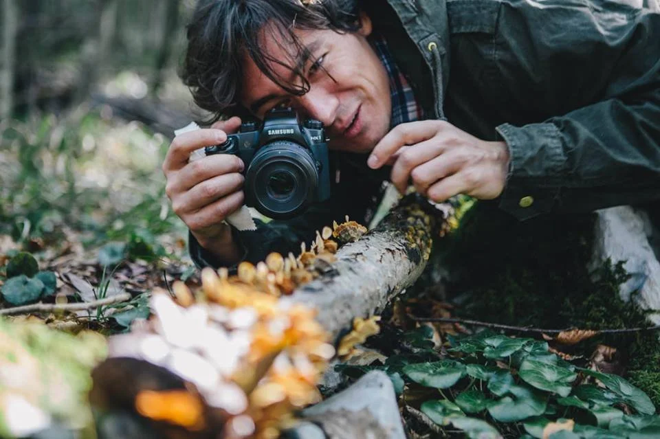 A man lying on the forest floor, taking a close-up photograph of mushrooms growing on a fallen log.