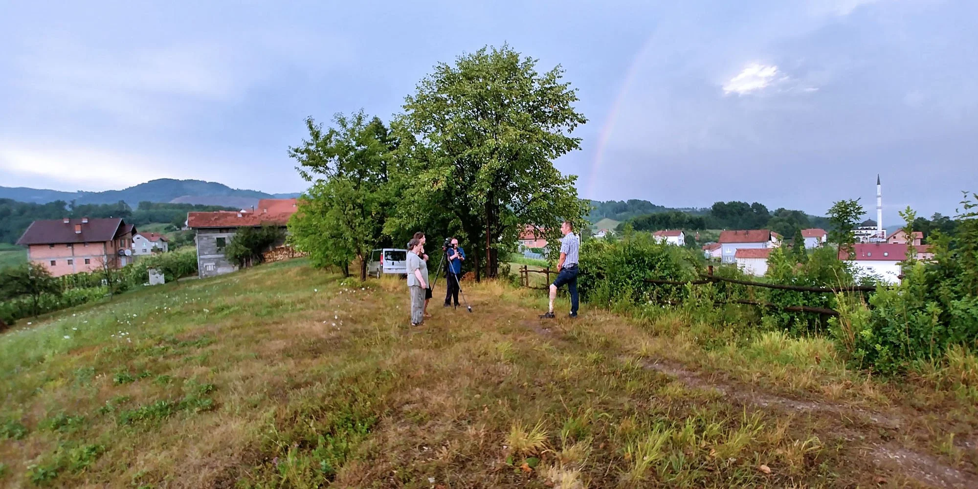 A group of people, including a woman, a man, and a cameraman, are gathered outdoors near a large tree on a grassy hill in a small town or village. The scene includes houses, a church steeple, and a rainbow in the background, with a cloudy sky overhea