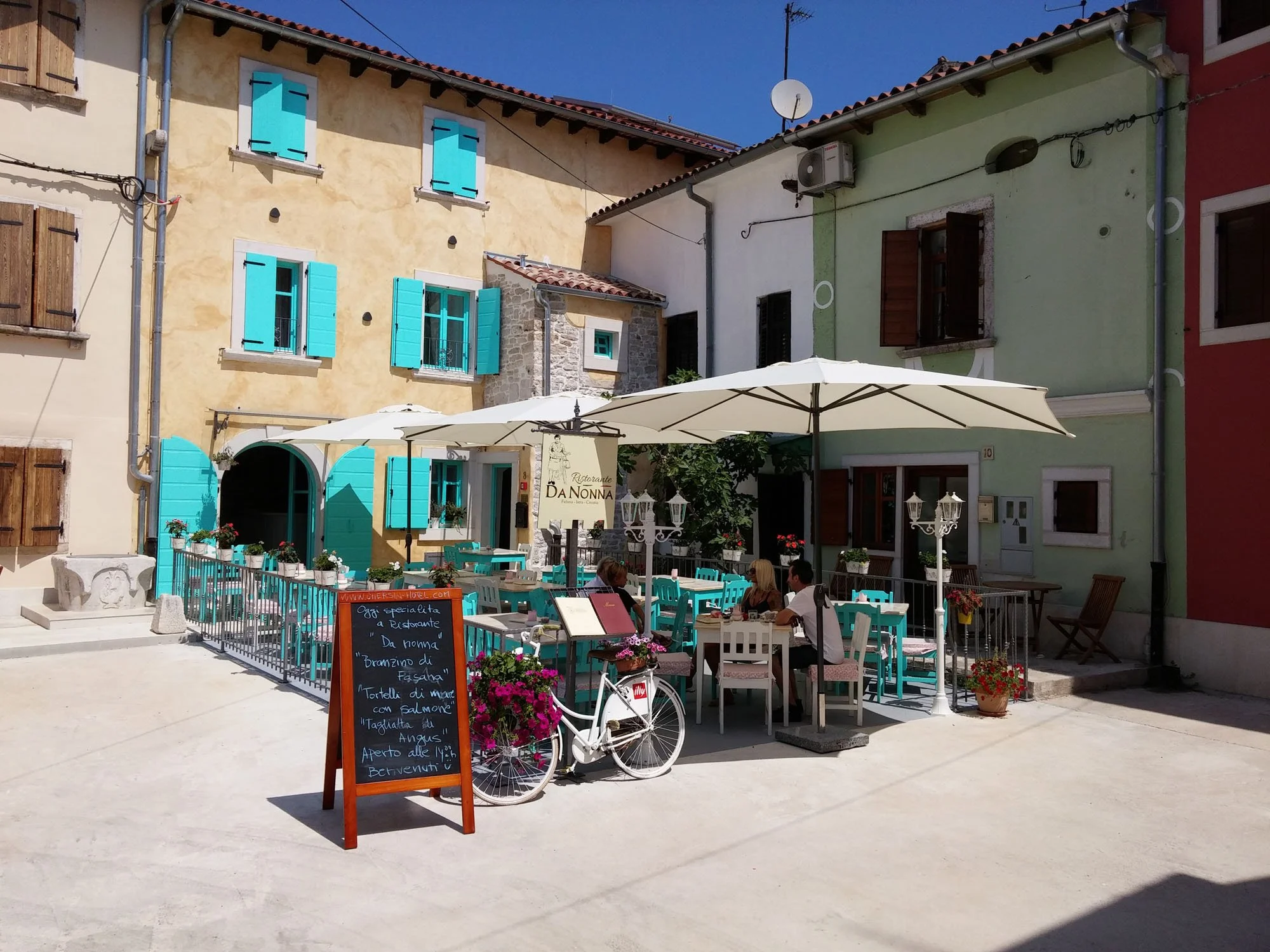 A small outdoor café with light blue chairs and white tables, shaded by large white umbrellas, in a European-style square with colorful buildings and outdoor seating.