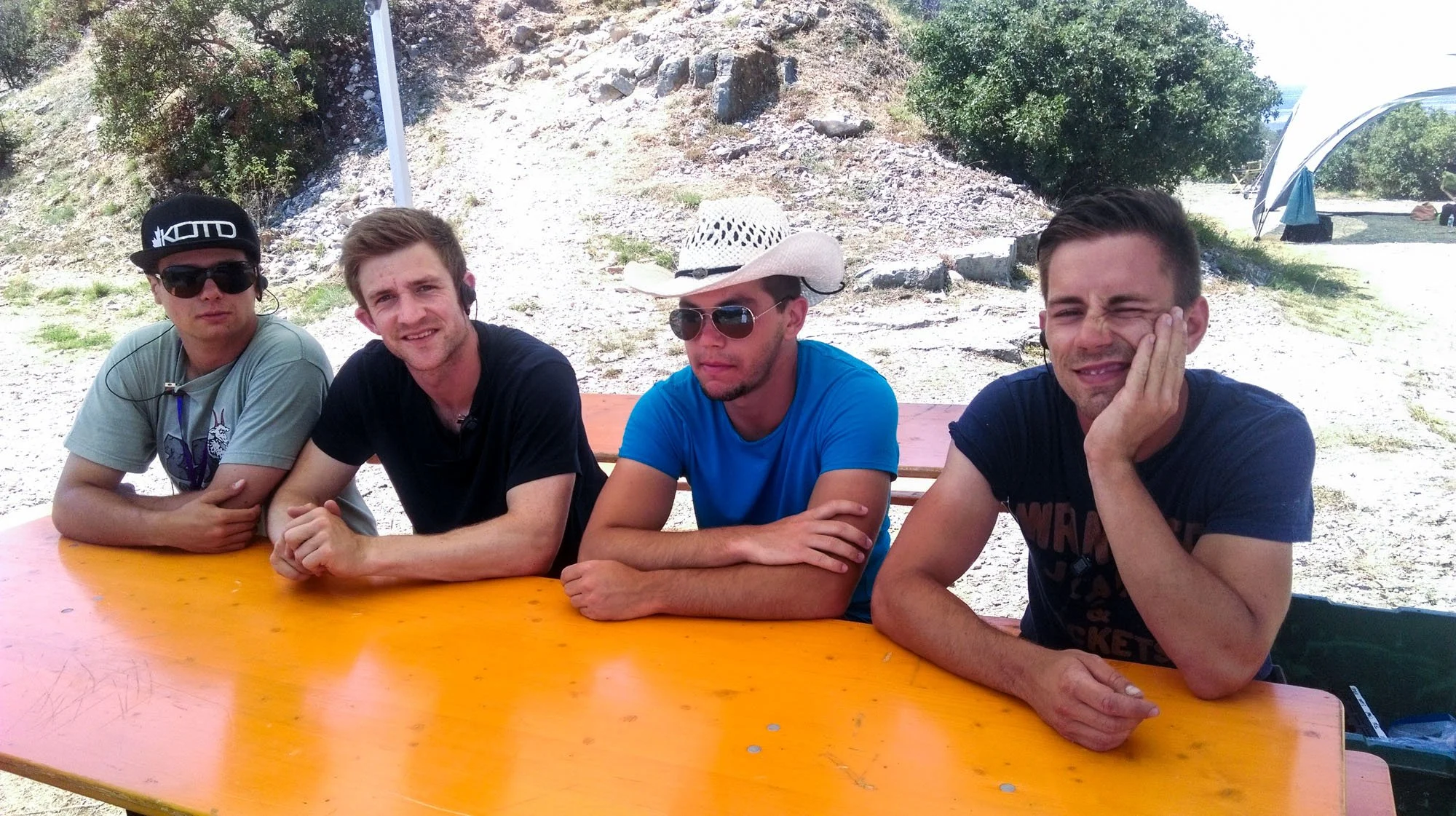 Four young men sitting at an orange picnic table outdoors on a sunny day, with trees, rocks, and a tent in the background.